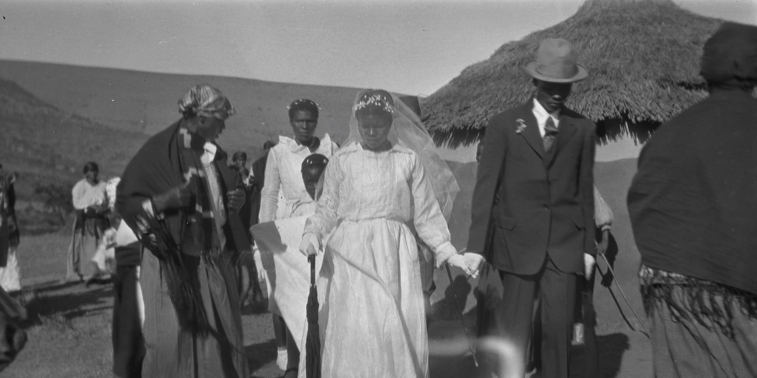 An old black-and-white photo shows a traditional rural setting, where an African couple walks hand-in-hand in formal western wedding attire, past villagers in more traditional attire.
