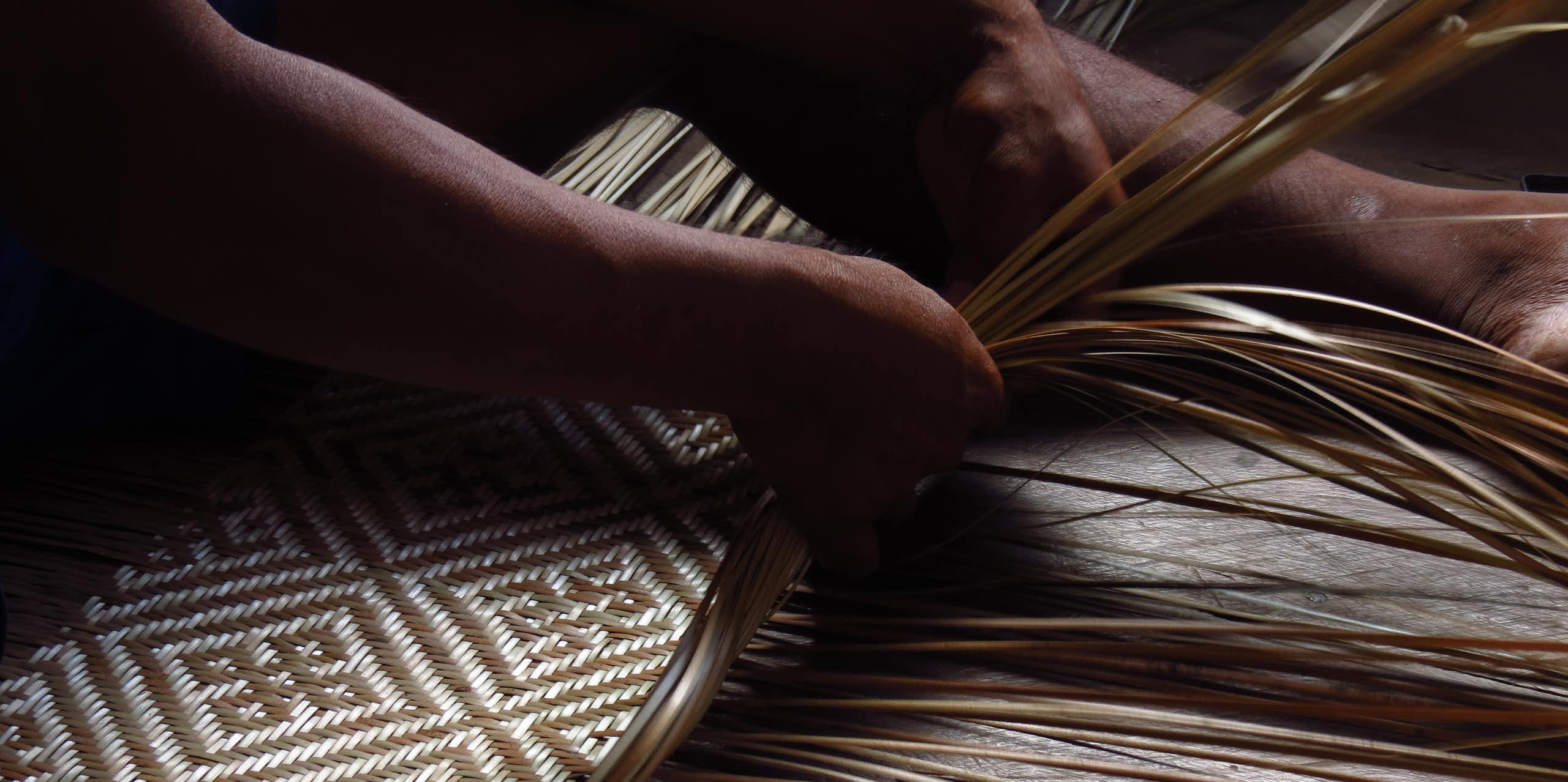 Foto mostra as mãos de um indígena tecendo fibras vegetais e criando um padrão geométrico de 2 cores, bege e marrom