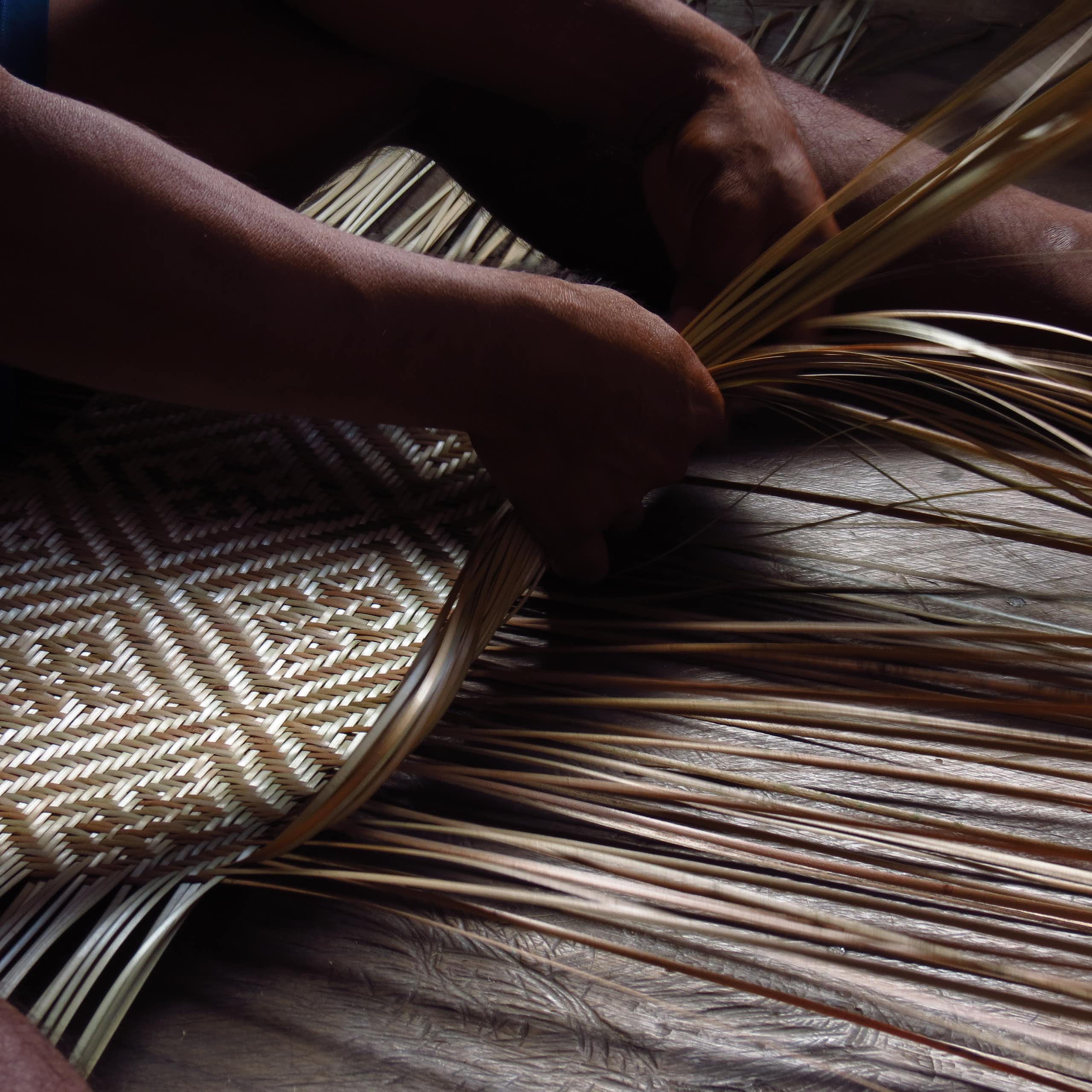 Foto mostra as mãos de um indígena tecendo fibras vegetais e criando um padrão geométrico de 2 cores, bege e marrom