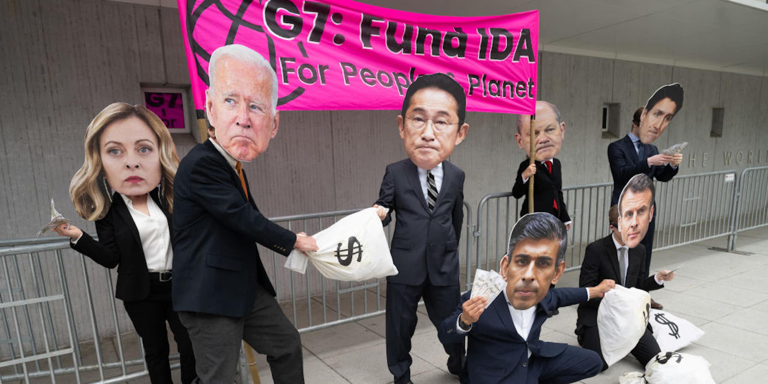 In front of a banner, people in suits carry bags labelled with dollar signs and wear large masks representing the faces of world leaders