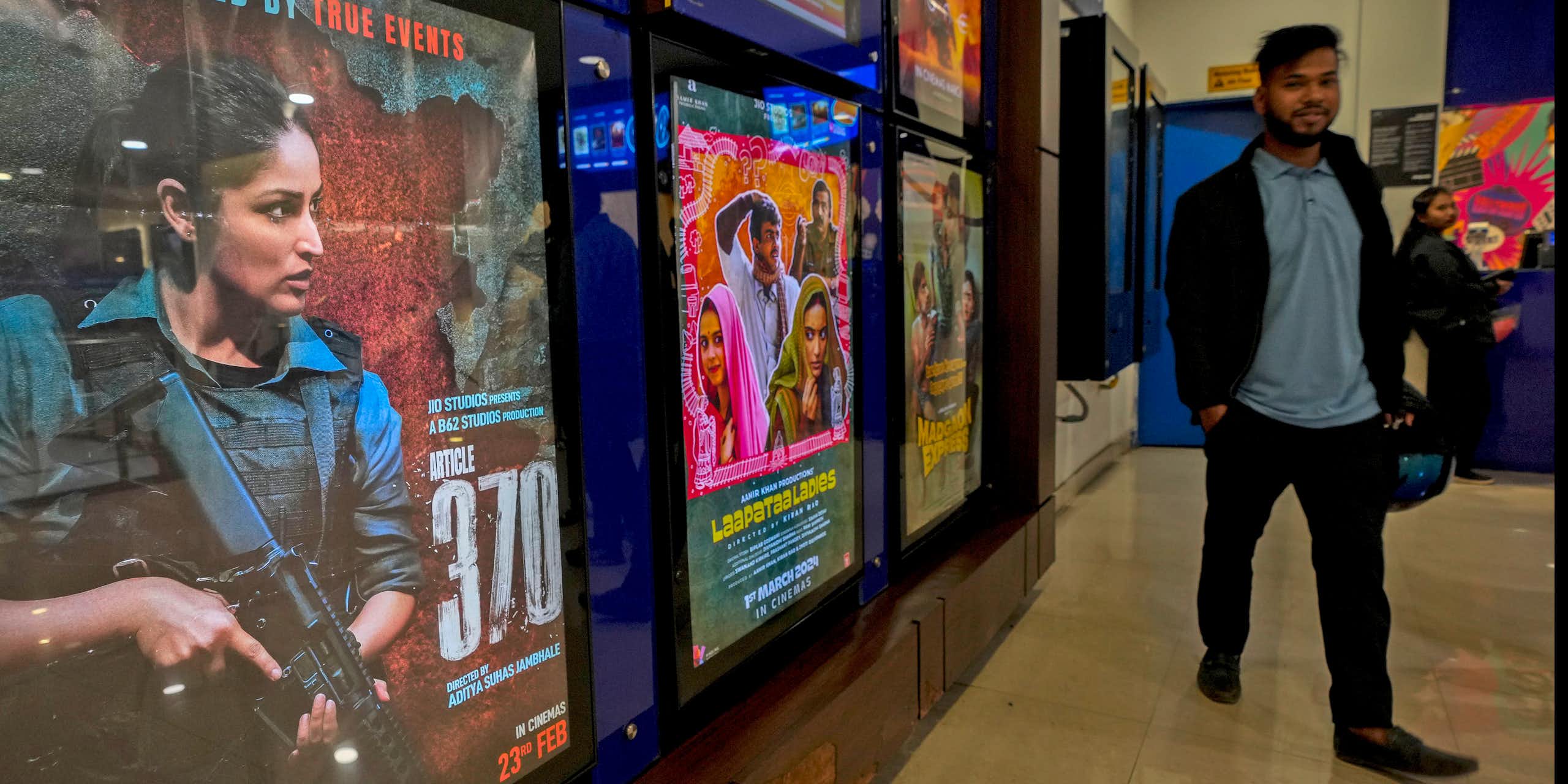 A movie poster shows a woman holding an automatic rifle. A man walks beside the film poster.