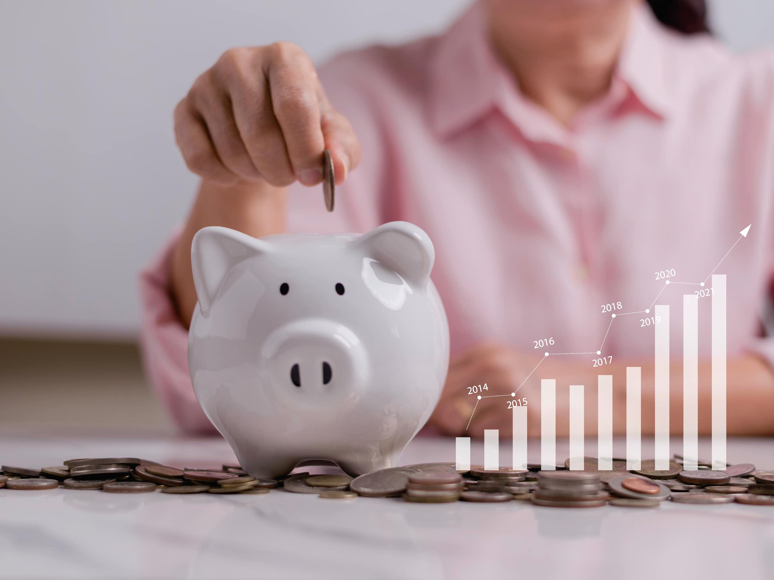 A person, seen from the neck down, puts a coin in a piggy bank. A visual of a bar graph increasing in value over time is seen beside it.