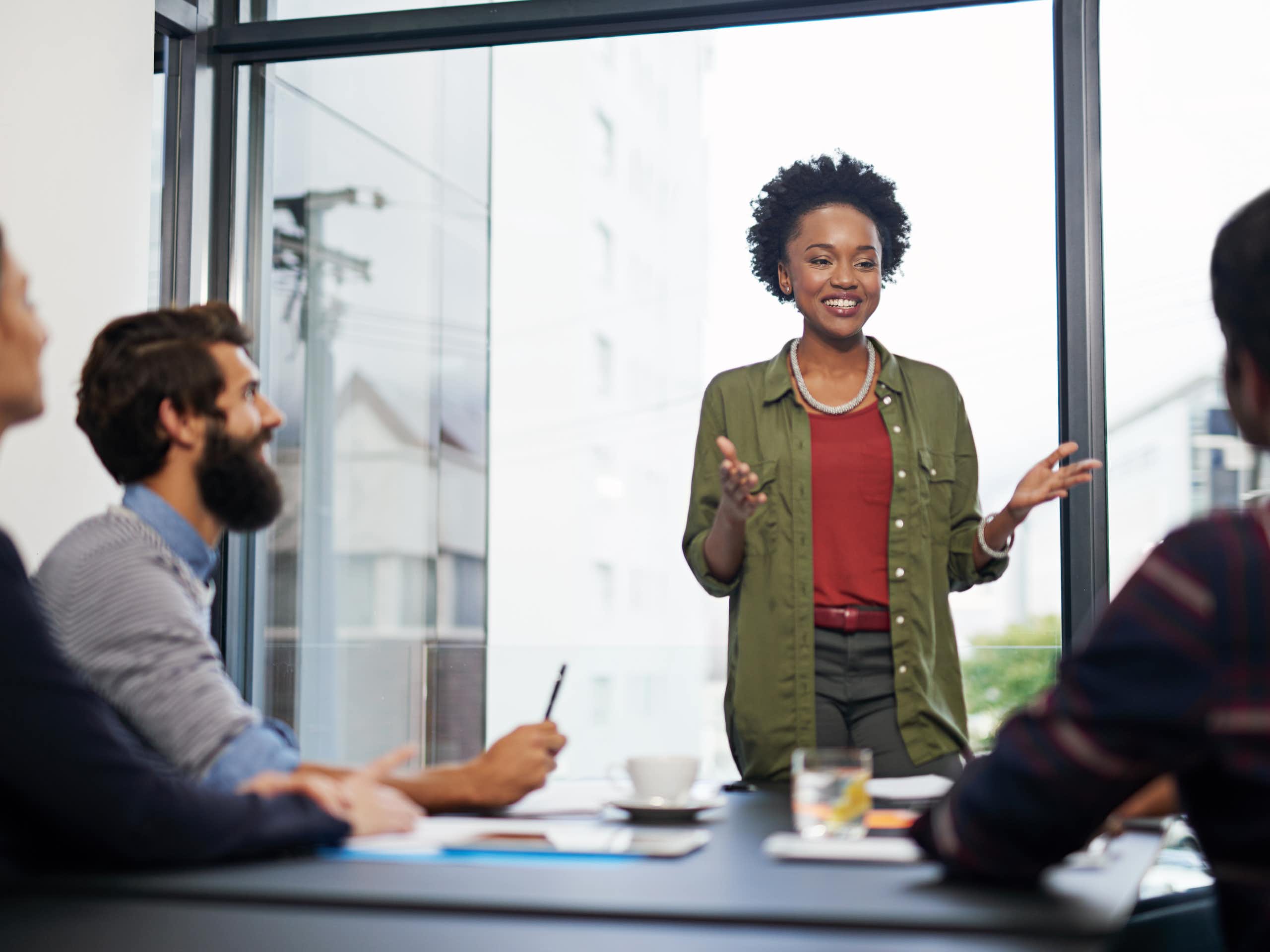 A young woman stands before a table of people in an office, gesturing with her hands while speaking to them