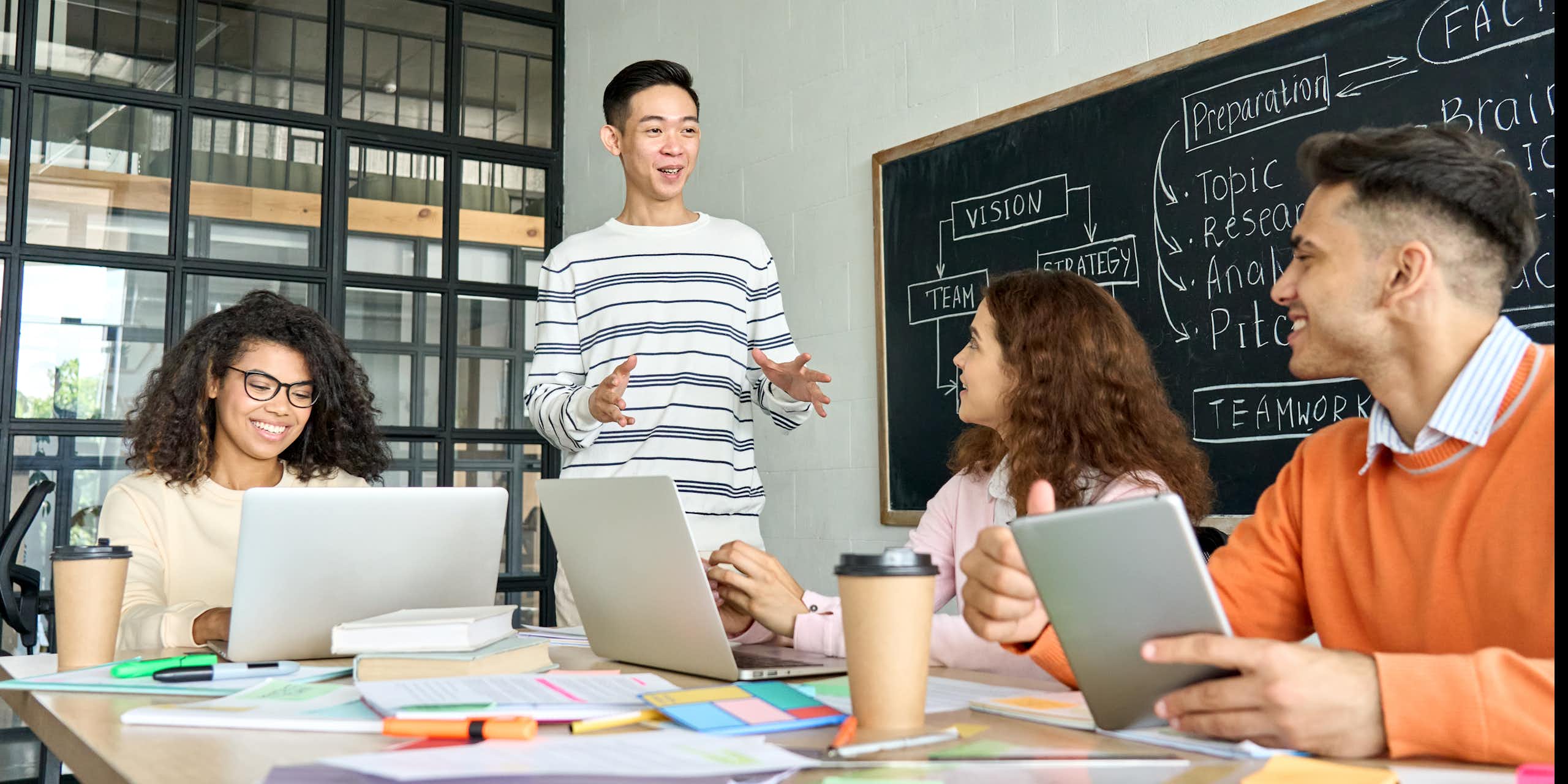 A young man stands while speaking to a group of peers who are sitting around a table