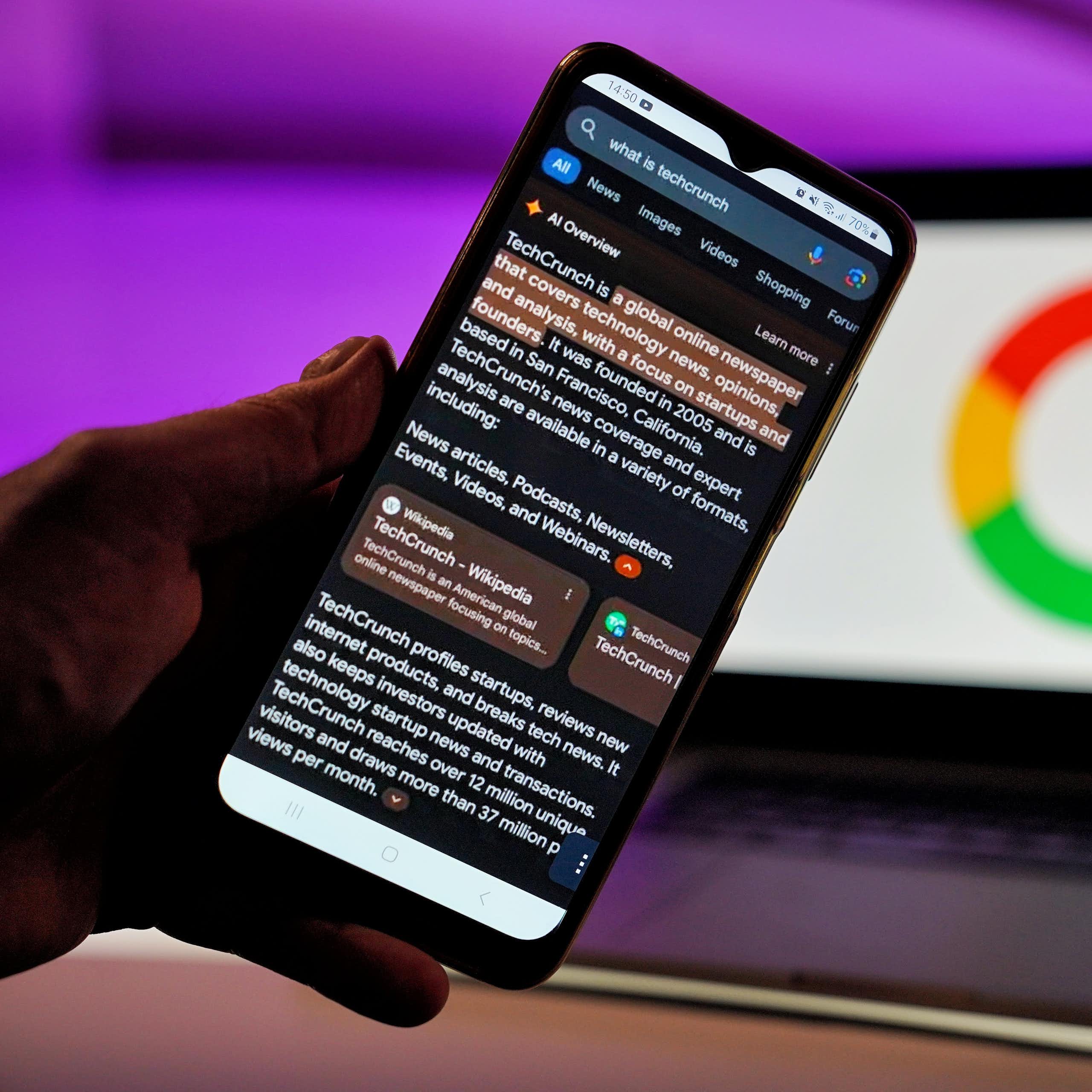 A smartphone help in front of an open laptop displaying the Google logo displays a Google search engine result with a section at the top of the search that says 'AI Overview' followed by an AI-generated response to a search
