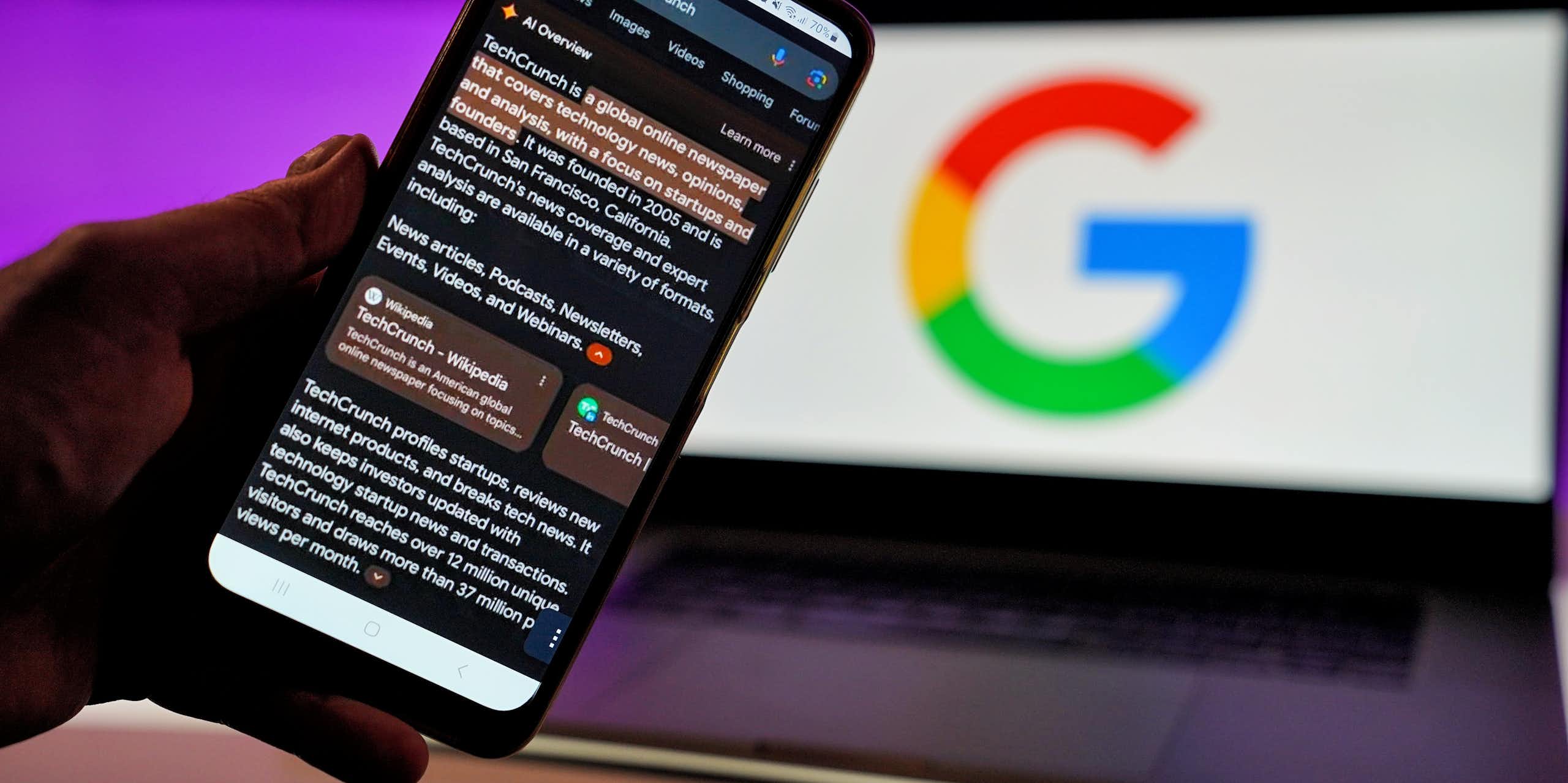 A smartphone help in front of an open laptop displaying the Google logo displays a Google search engine result with a section at the top of the search that says 'AI Overview' followed by an AI-generated response to a search