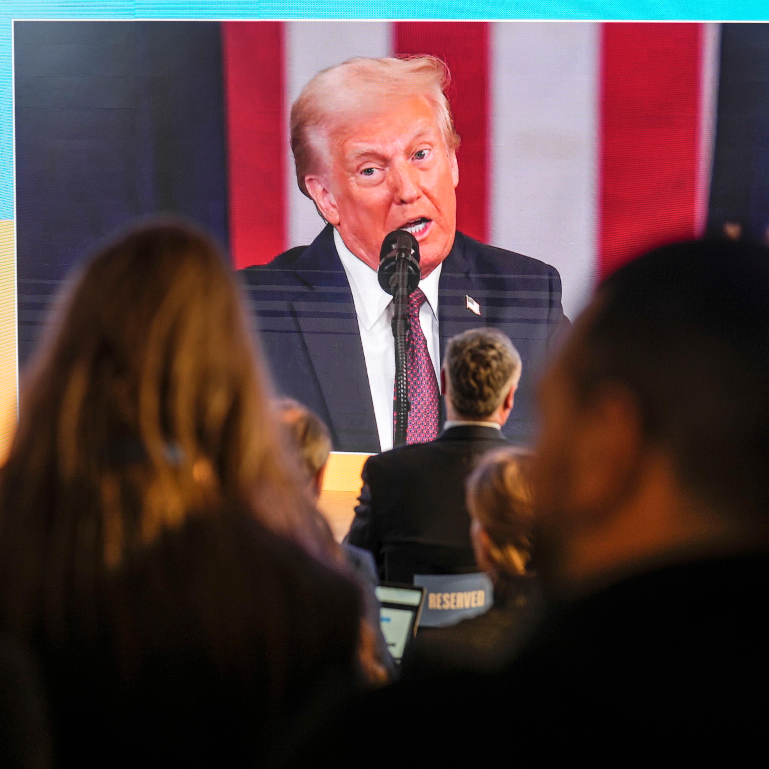 A man with fluffy grey-blond hair appears on a jumbo screen against the backdrop of a giant blue and yellow flag as people watch him speak.