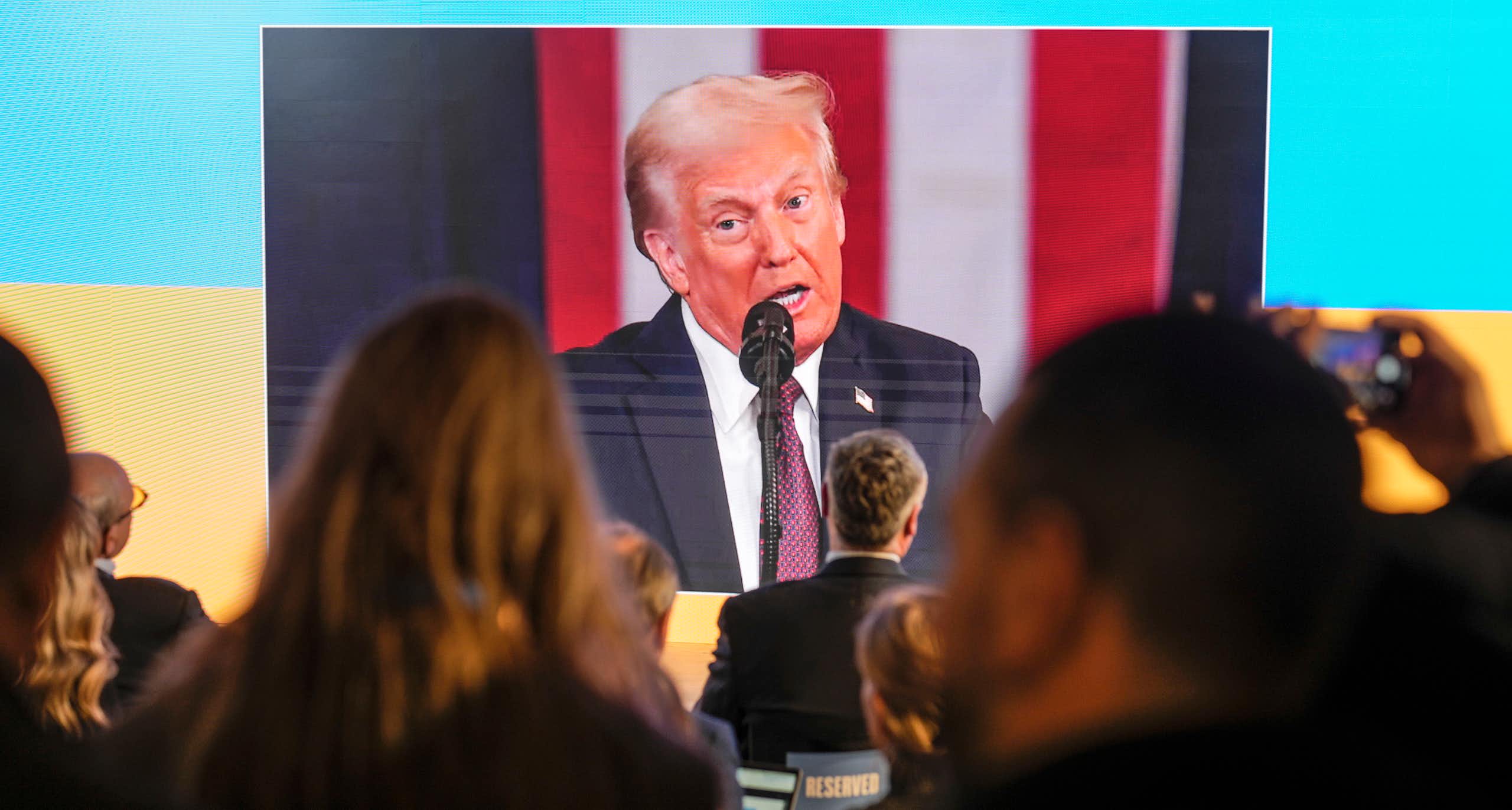 A man with fluffy grey-blond hair appears on a jumbo screen against the backdrop of a giant blue and yellow flag as people watch him speak.