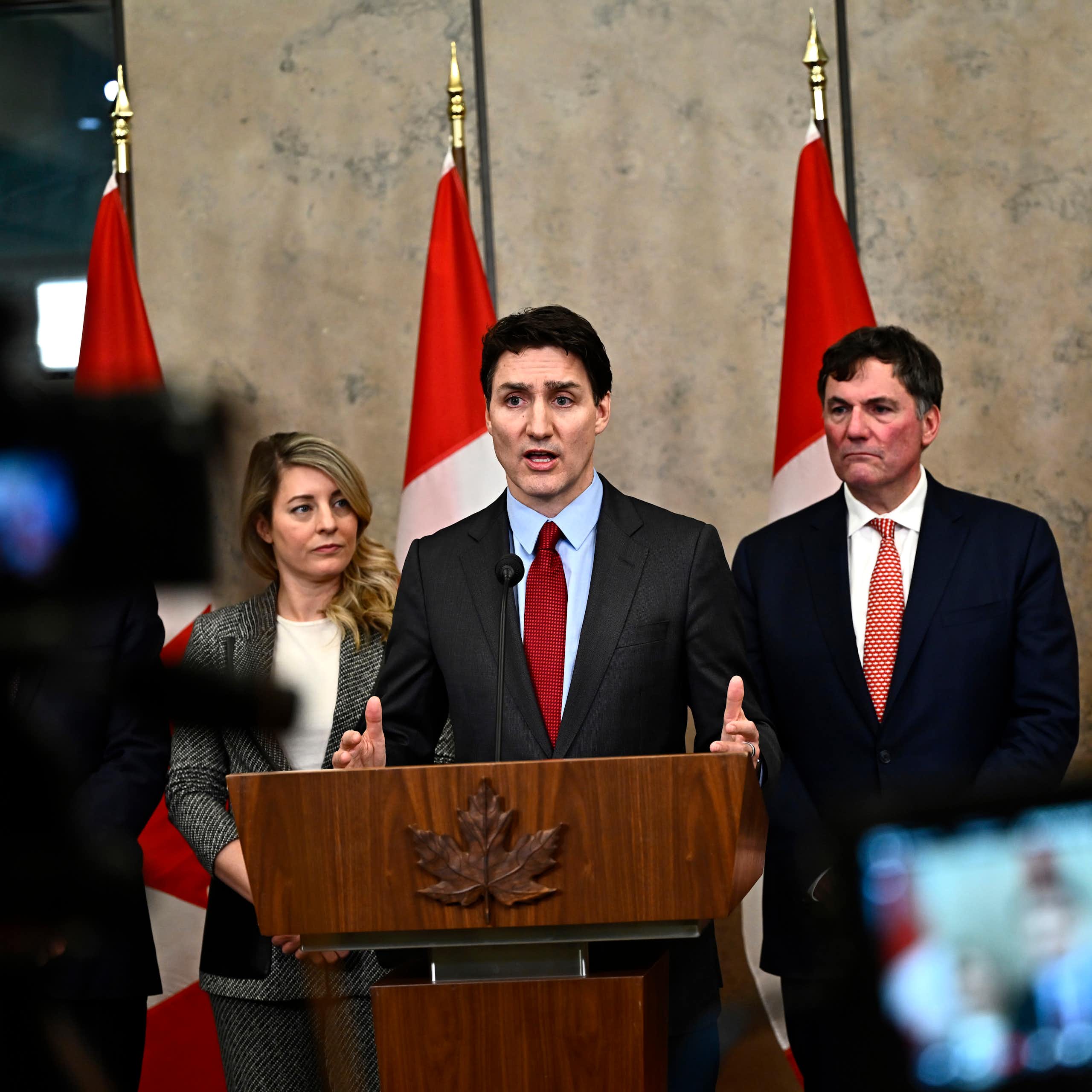 Un homme blanc en costume s'exprime depuis un podium, avec des drapeaux canadiens derrière lui. Il est entouré d'une femme blanche et d'un homme blanc.