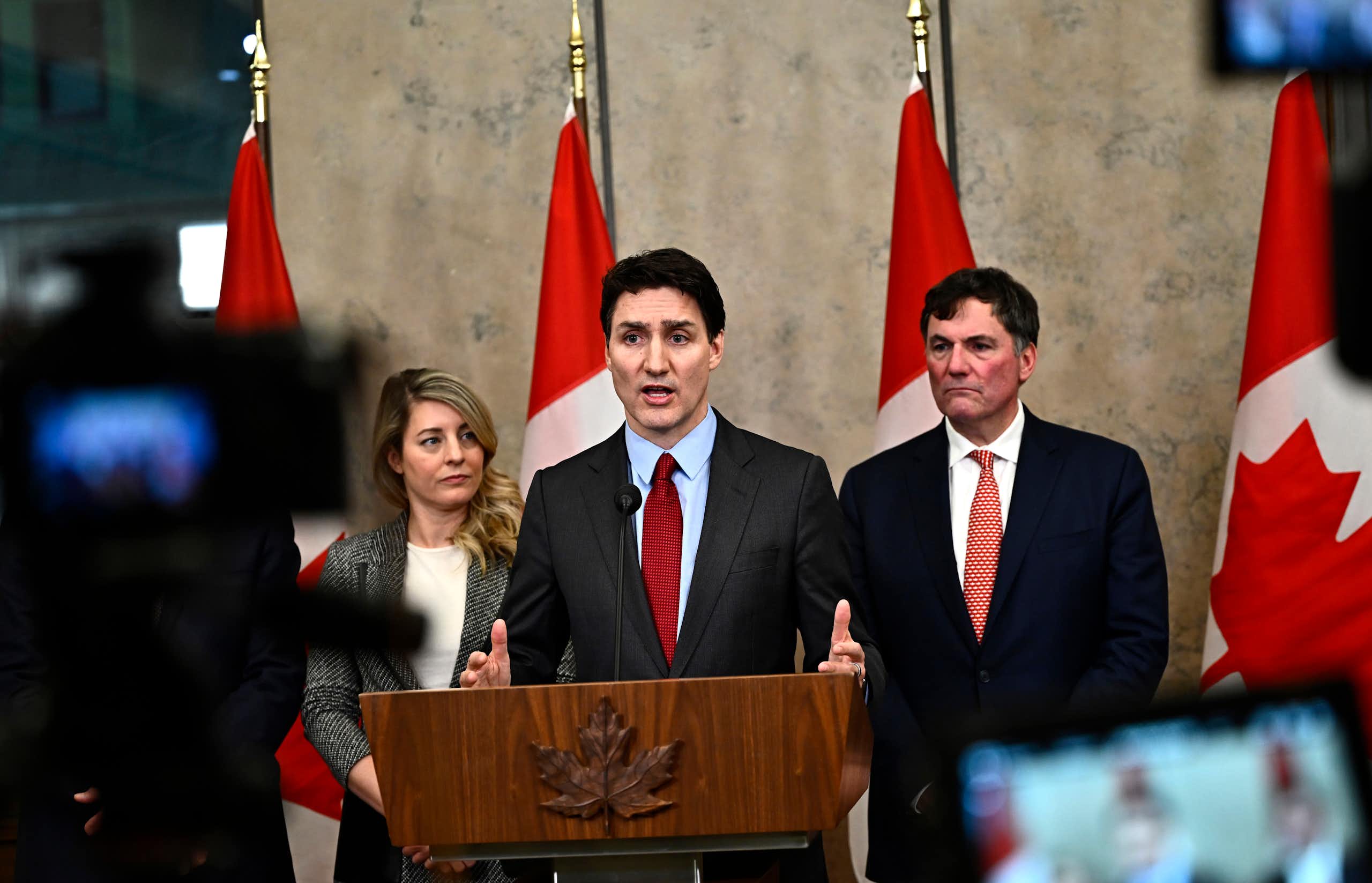 Un homme blanc en costume s'exprime depuis un podium, avec des drapeaux canadiens derrière lui. Il est entouré d'une femme blanche et d'un homme blanc.