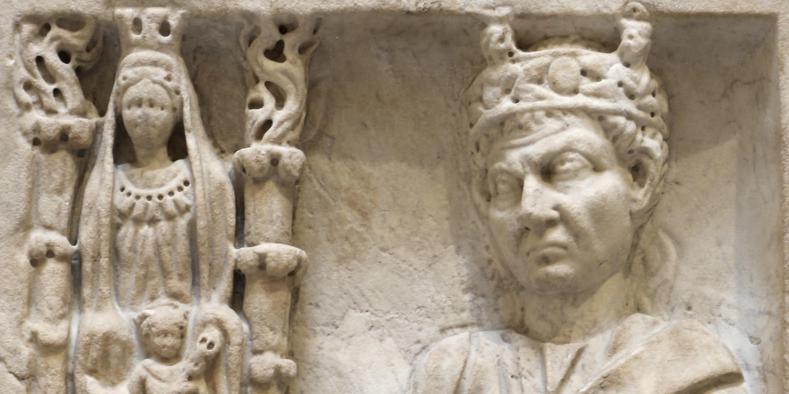 Um relevo de mármore com várias figuras gravadas: ao lado, uma coluna com uma mulher adornada e outra acompanhante. Ao lado deles está um homem com uma coroa, com uma das mãos em um pedestal.