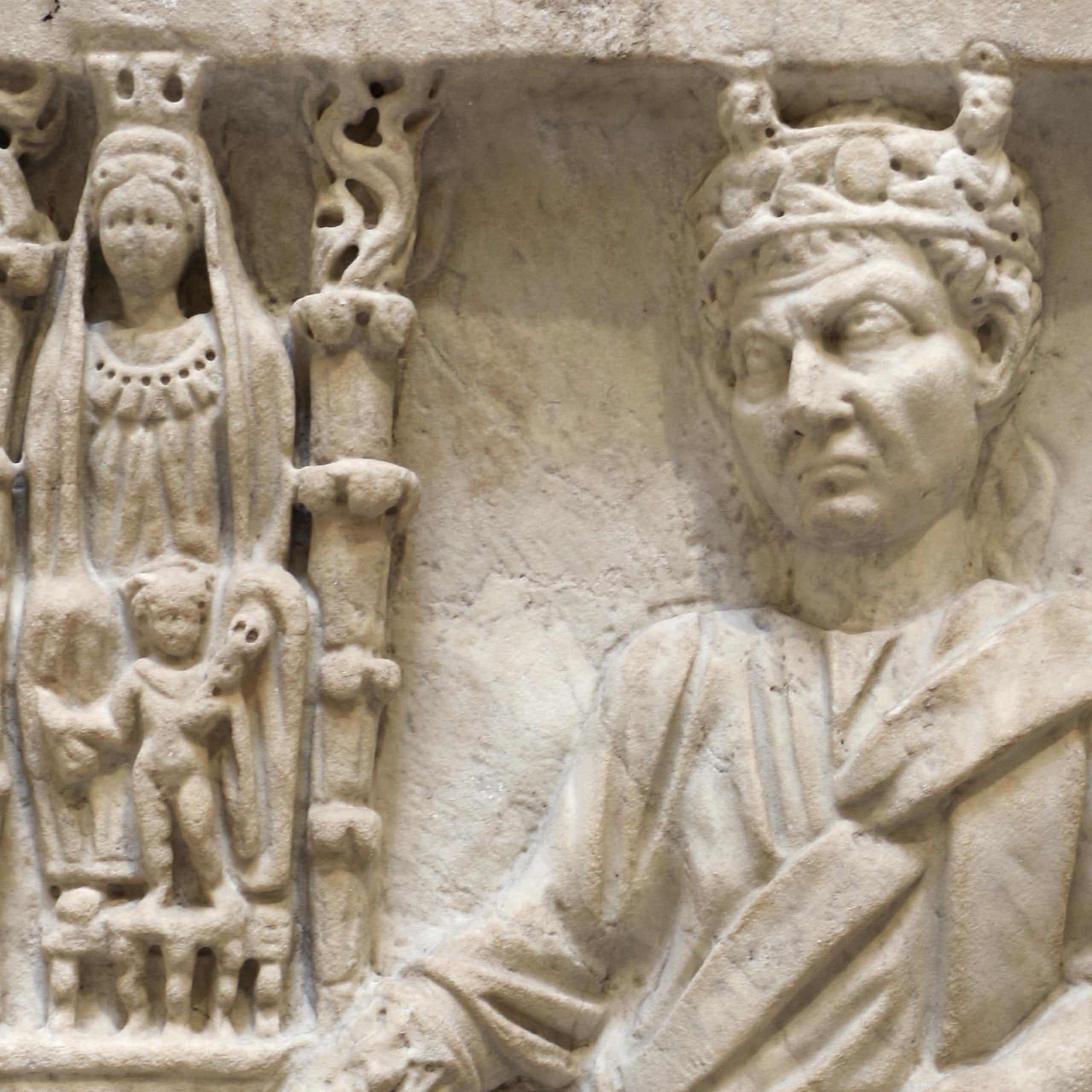 Um relevo de mármore com várias figuras gravadas: ao lado, uma coluna com uma mulher adornada e outra acompanhante. Ao lado deles está um homem com uma coroa, com uma das mãos em um pedestal.