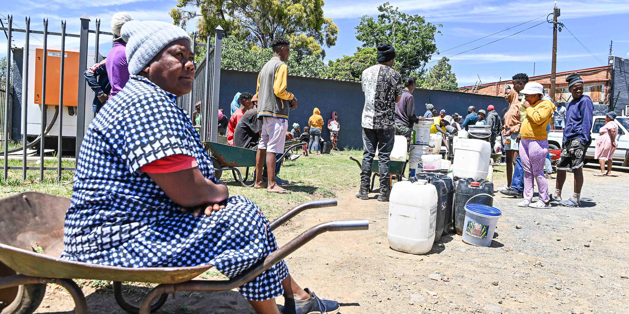 A woman sits on a wheelbarrow at the end of a queue of people lining up with buckets and containers for water
