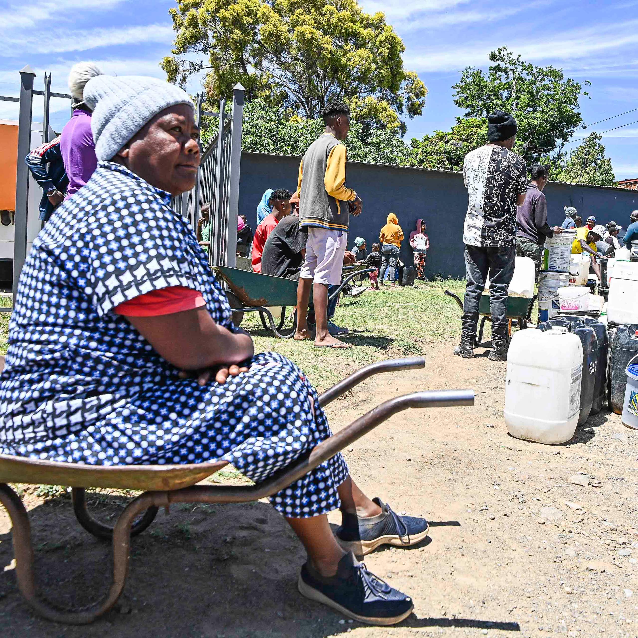 A woman sits on a wheelbarrow at the end of a queue of people lining up with buckets and containers for water