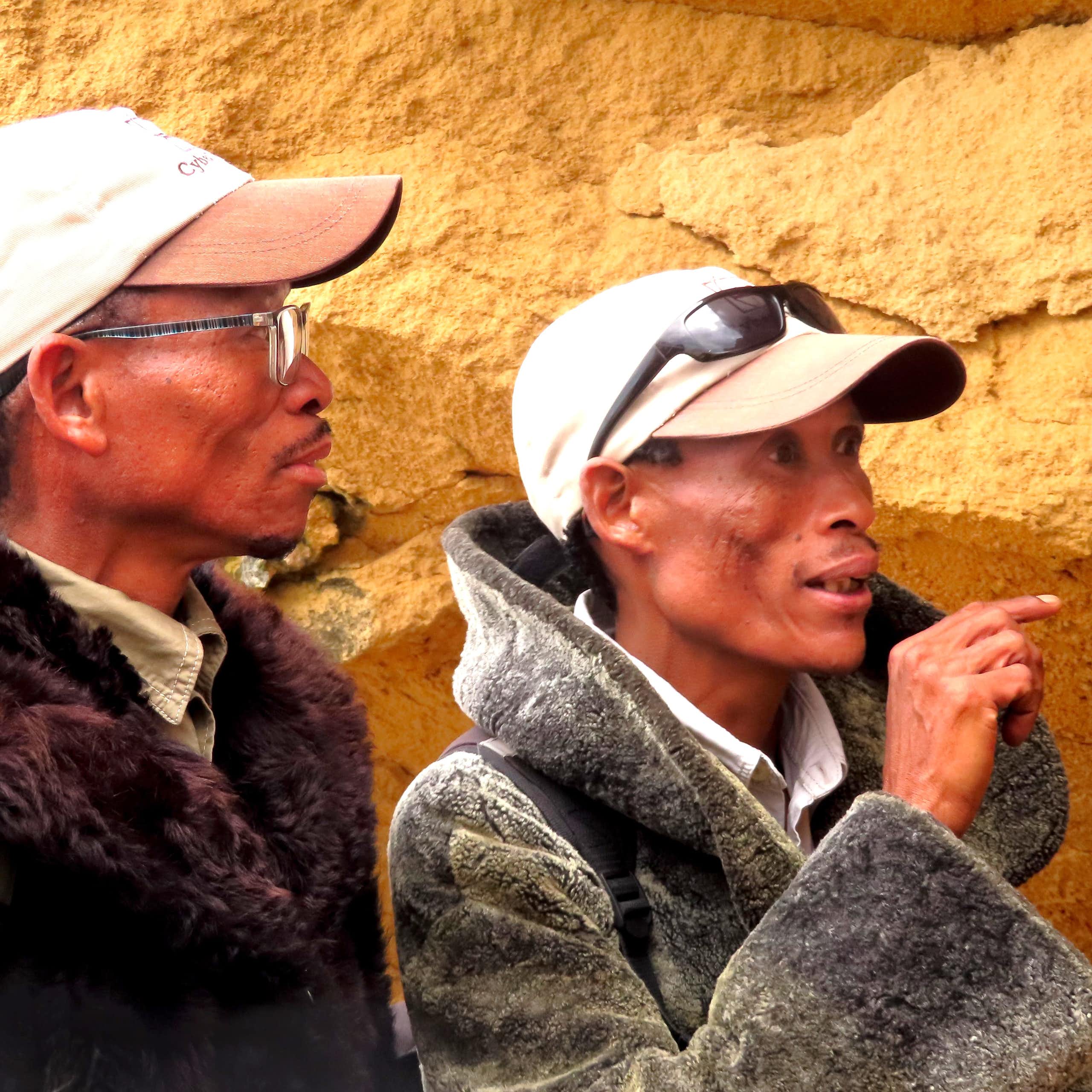 Two men wearing caps and warm jackets stand in front of a rocky wall. One of them is pointing ahead.