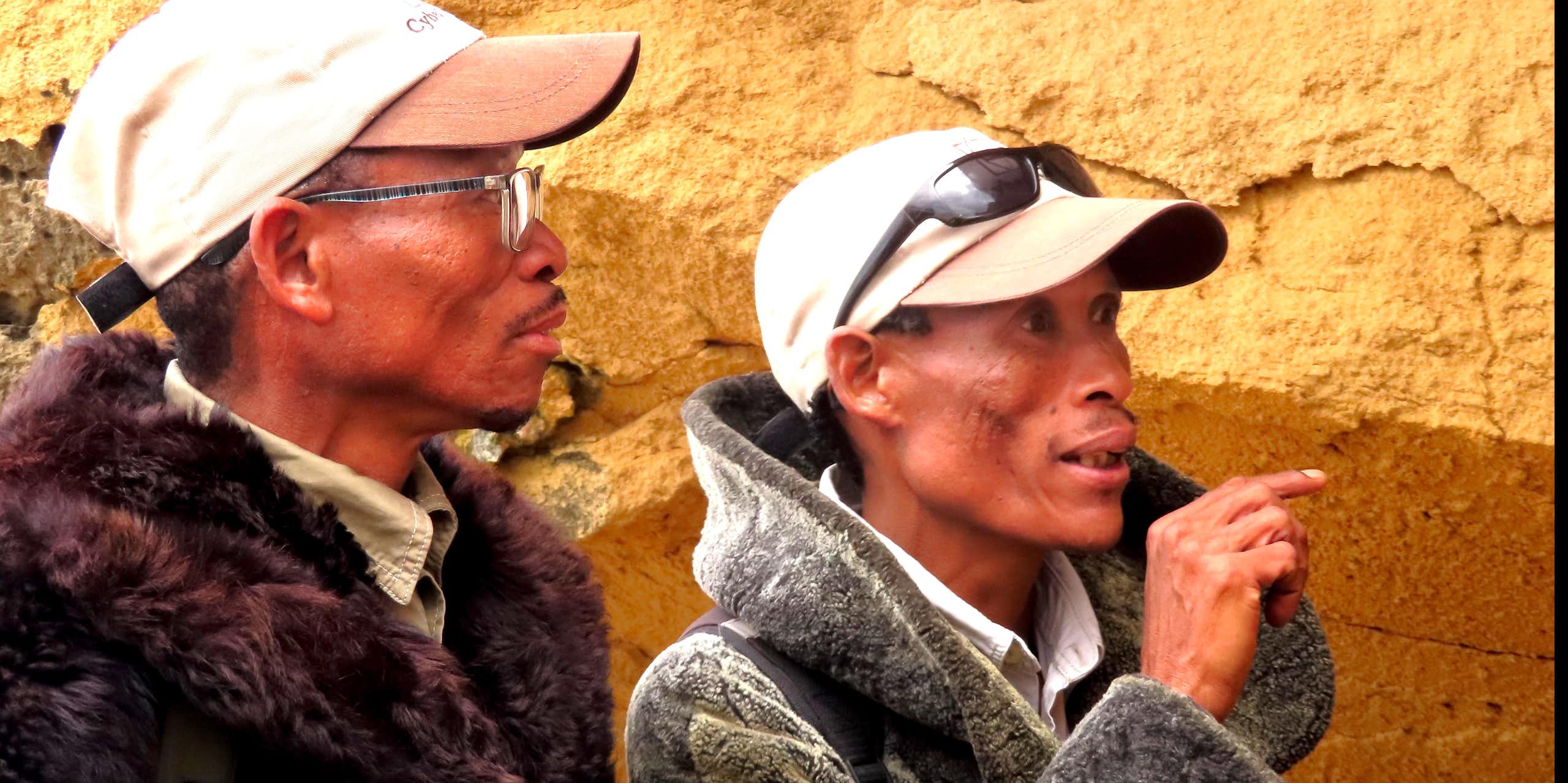 Two men wearing caps and warm jackets stand in front of a rocky wall. One of them is pointing ahead.