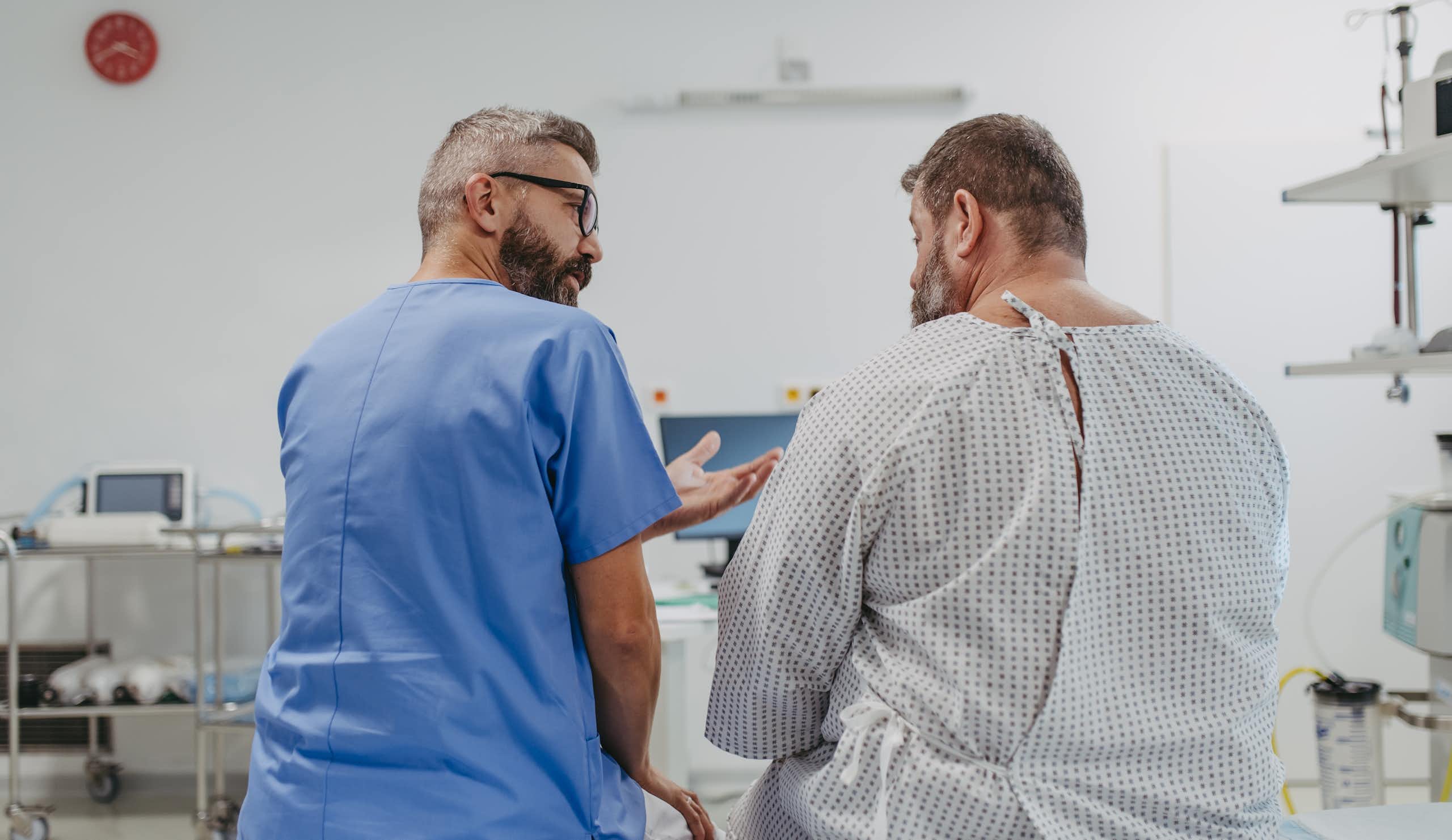 A doctor in blue scrubs sitting with an overweight man in a medical gown, seem from behind