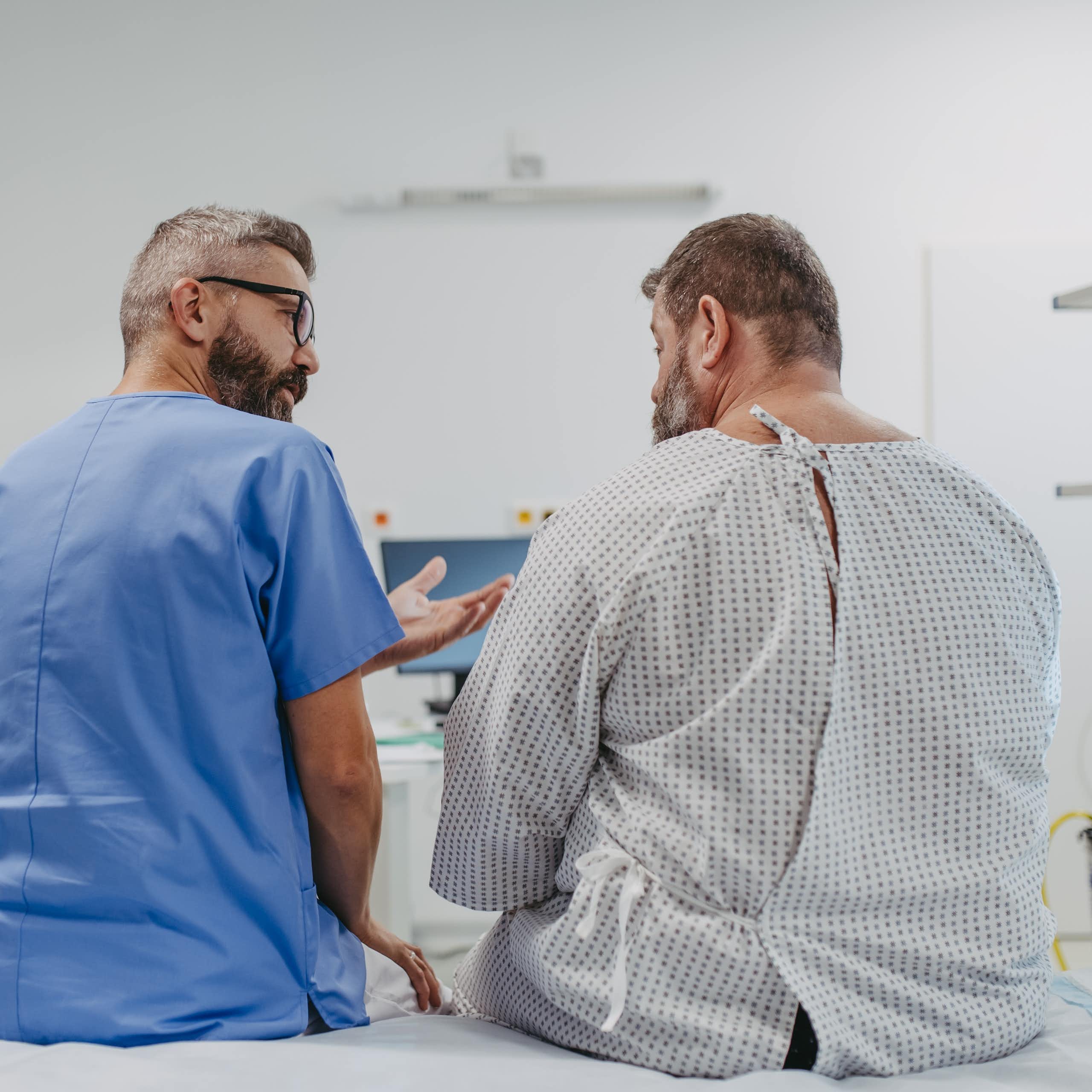 A doctor in blue scrubs sitting with an overweight man in a medical gown, seem from behind