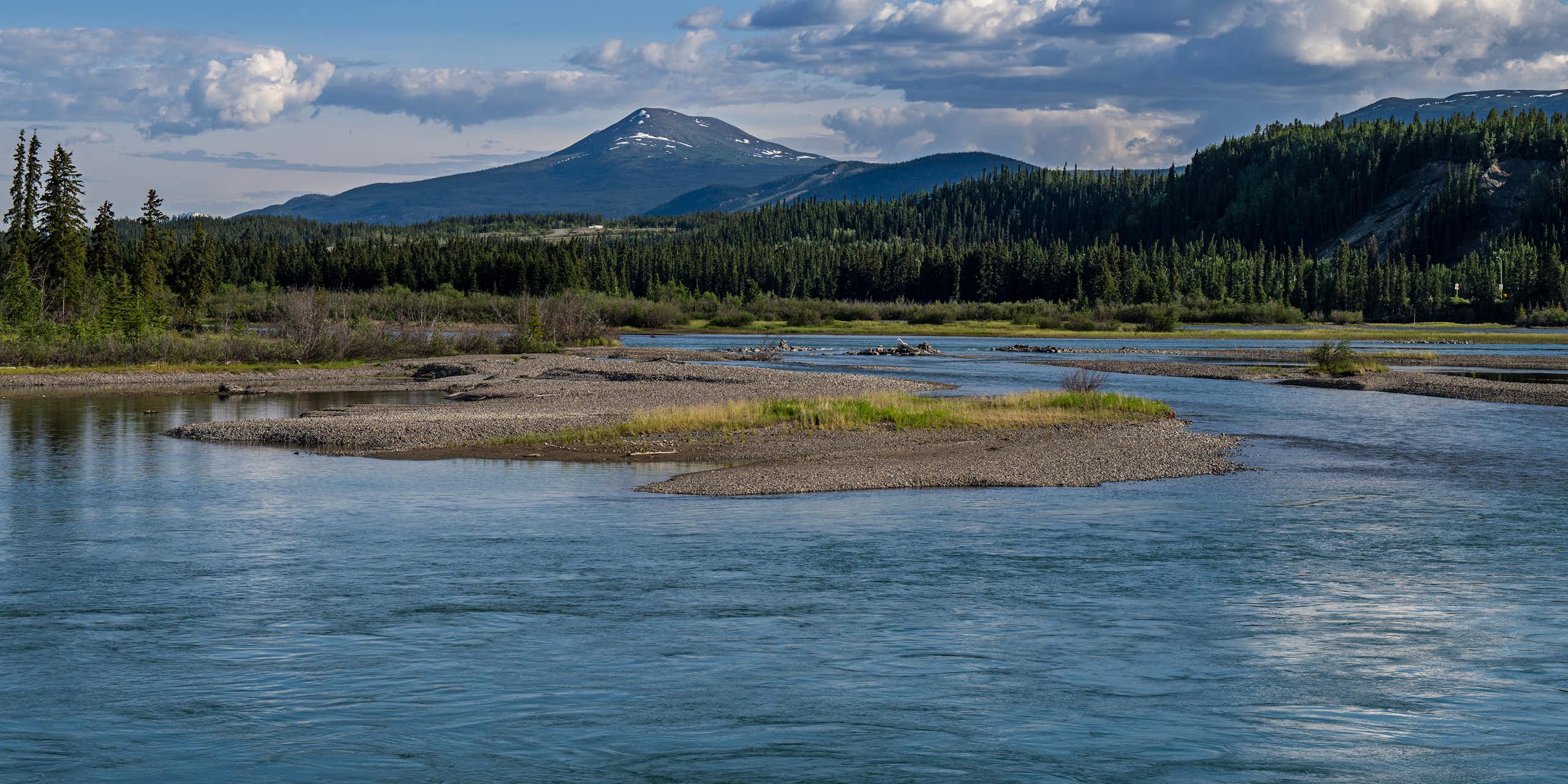 A wide blue river with mountains and pine trees surrounding it.
