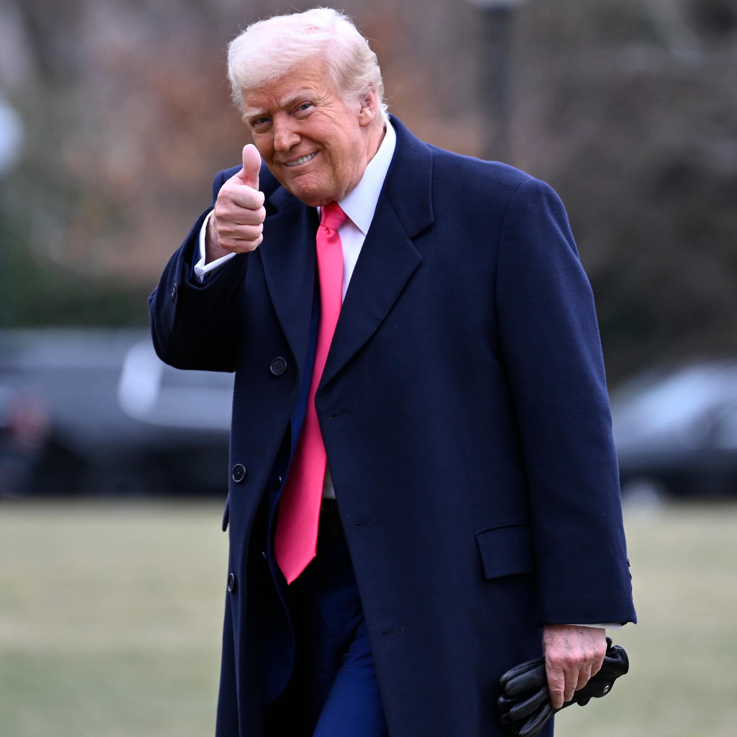 An old man with orange-tinged skin and whiteish hair wearing a dark suit and red tie gives a thumb's up to the camera