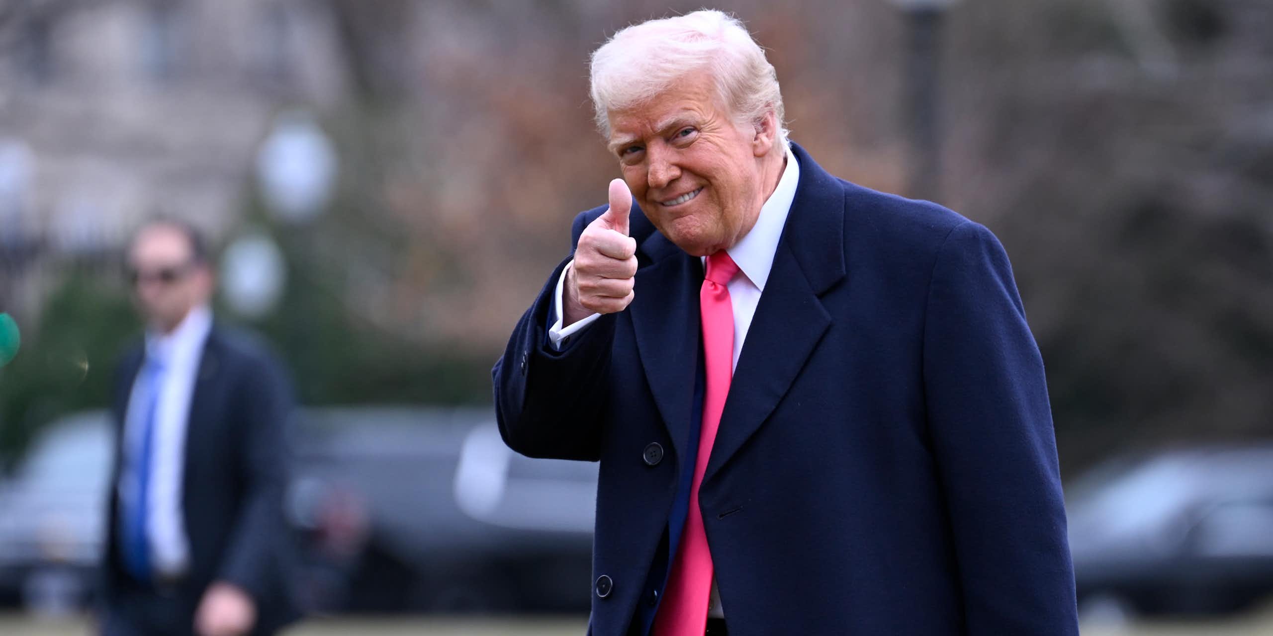 An old man with orange-tinged skin and whiteish hair wearing a dark suit and red tie gives a thumb's up to the camera