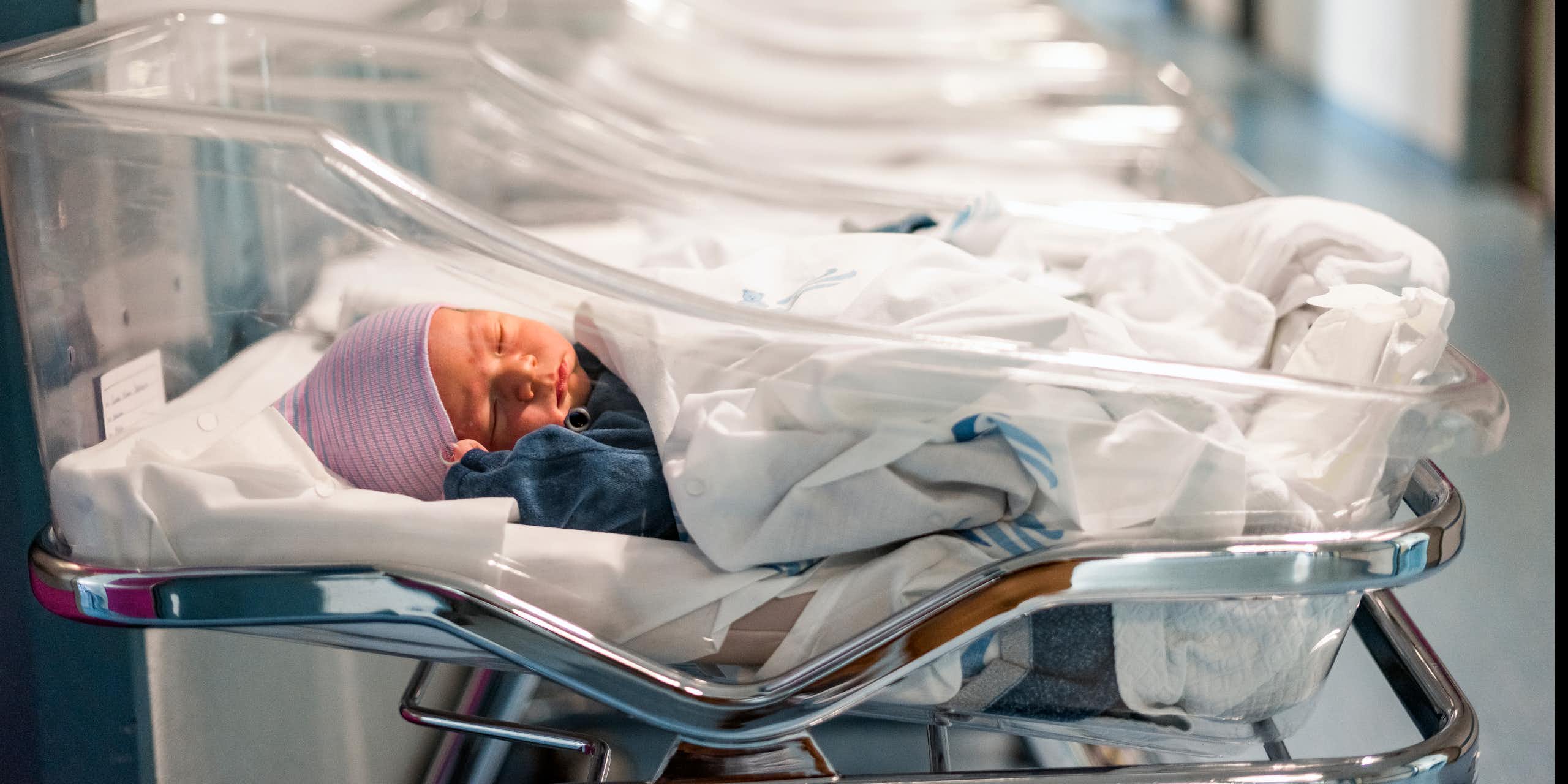 A newborn baby wearing a pink hat lays in a clear plastic bassinet in a hospital. A row of similar, empty bassinets are arranged alongside it.