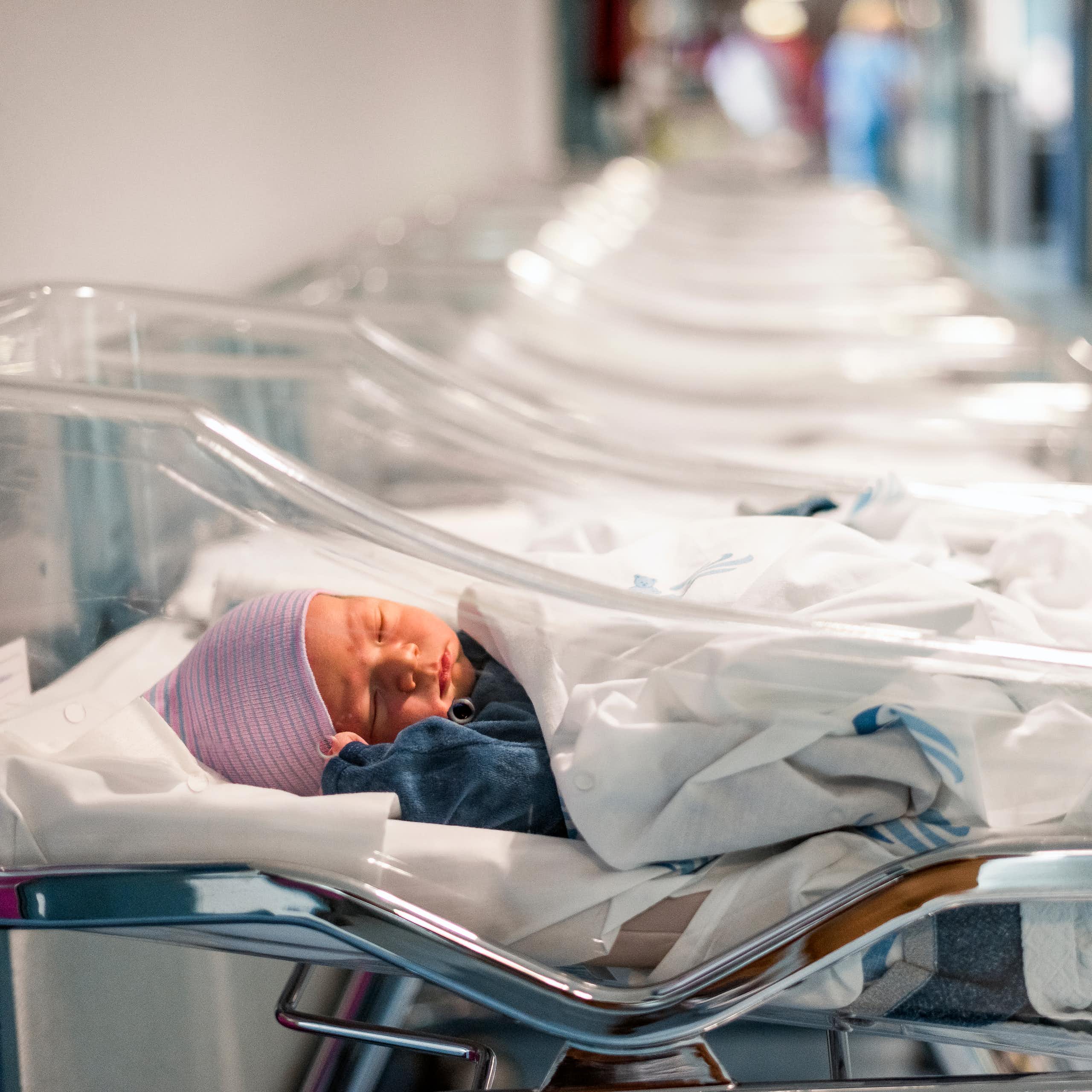 Dans un hôpital, un nouveau-né coiffé d'un bonnet rose est allongé dans un couffin en plastique transparent. Une rangée de berceaux similaires et vides sont disposés à côté de lui.