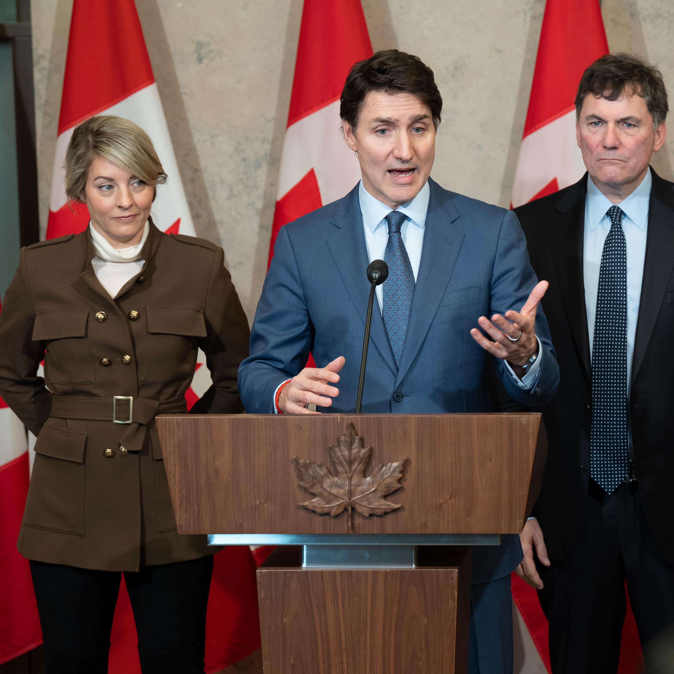 A white man with brown hair in a suit speaks from behind a podium. Behind him stand a white woman and two white men, all in suits