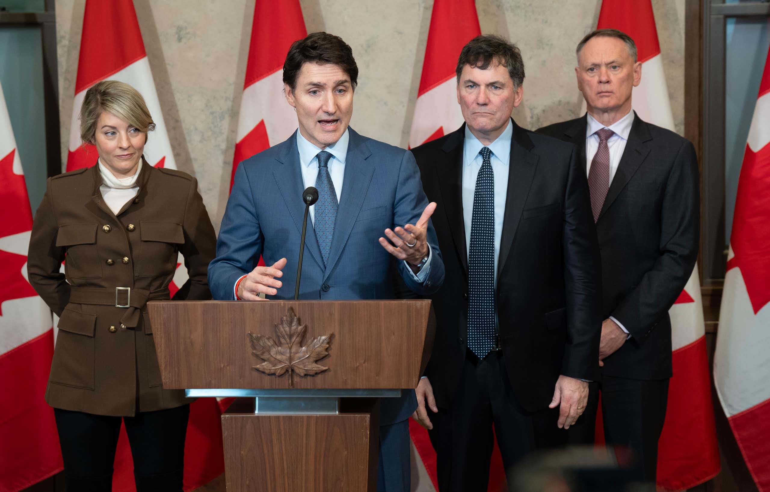A white man with brown hair in a suit speaks from behind a podium. Behind him stand a white woman and two white men, all in suits