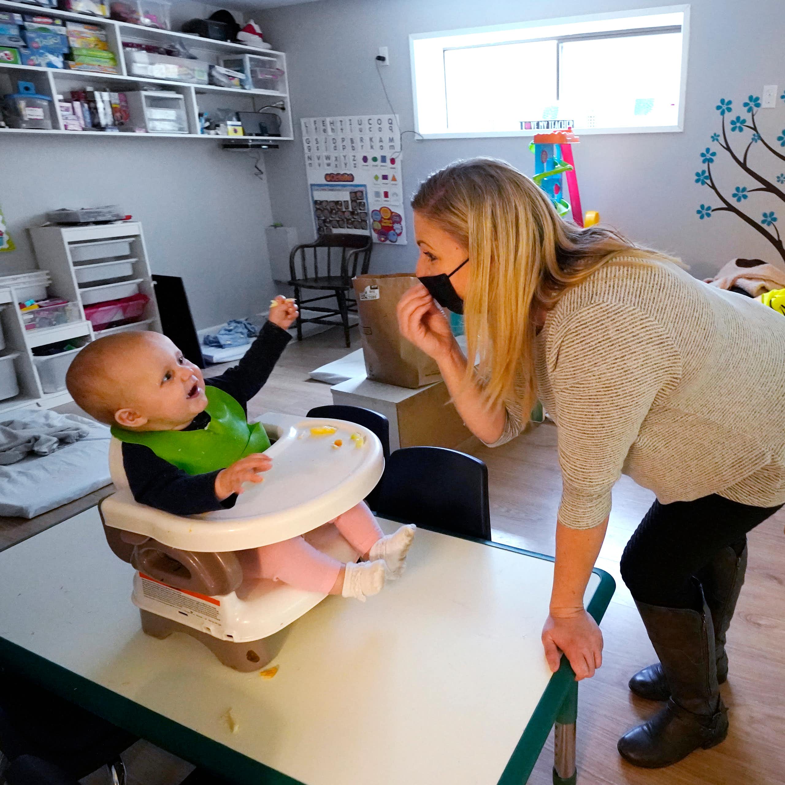 A baby sitting in a high chair reaches for their carer who is wearing a black COVID mask.  