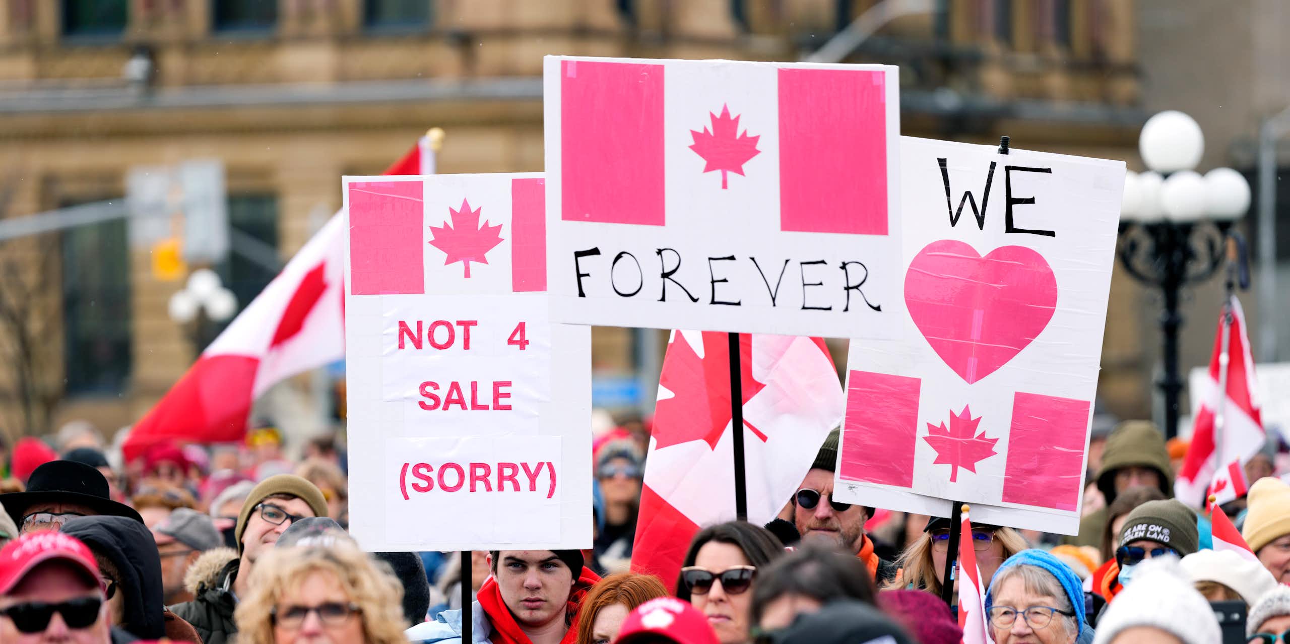 A crowd of protestors. Some of the people hold protest signs that state "Canada not 4 sale," "Canada Forever" and "We love Canada".