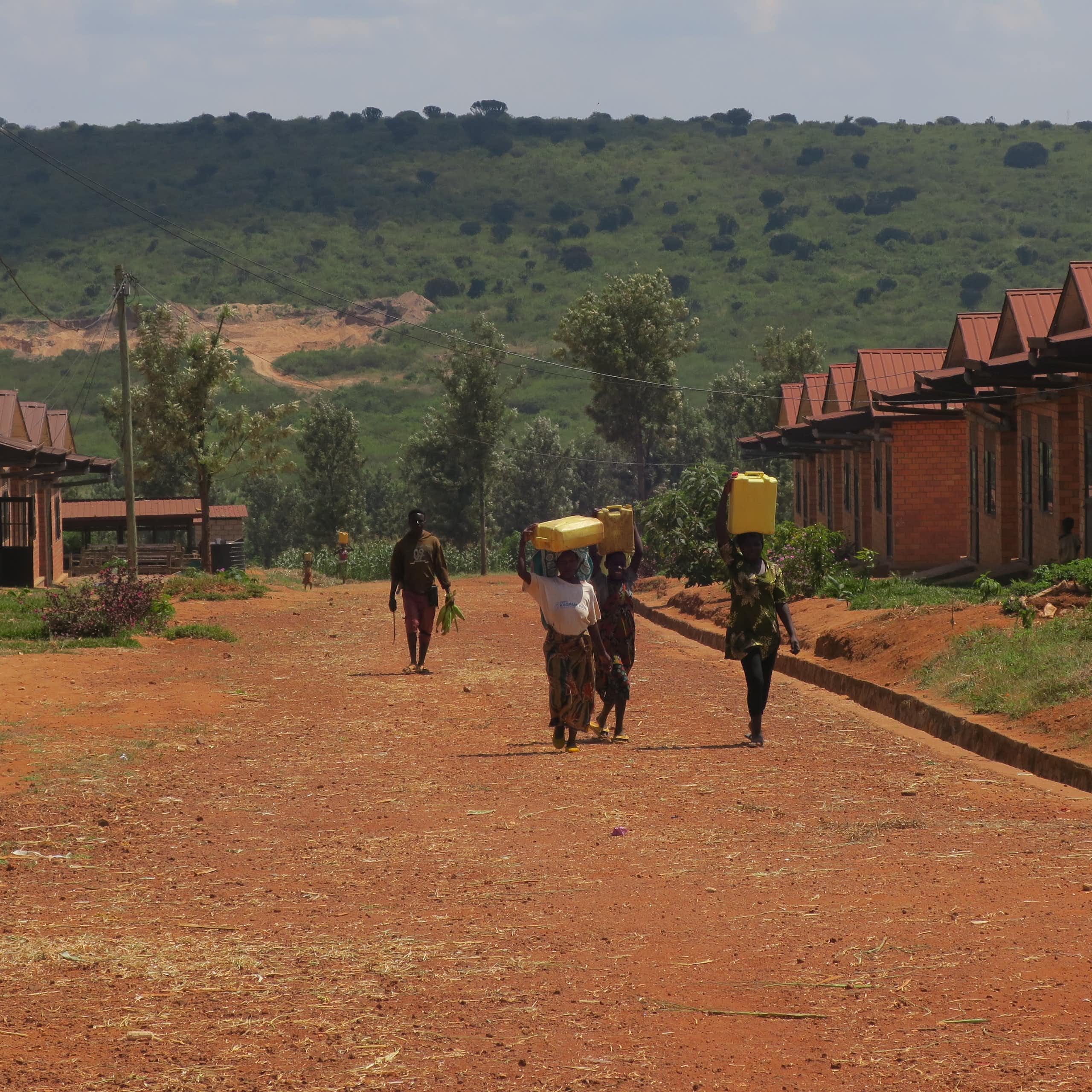 A dusty red road with two rows of houses on either side and people walking with 20 litre water buckets on their heads