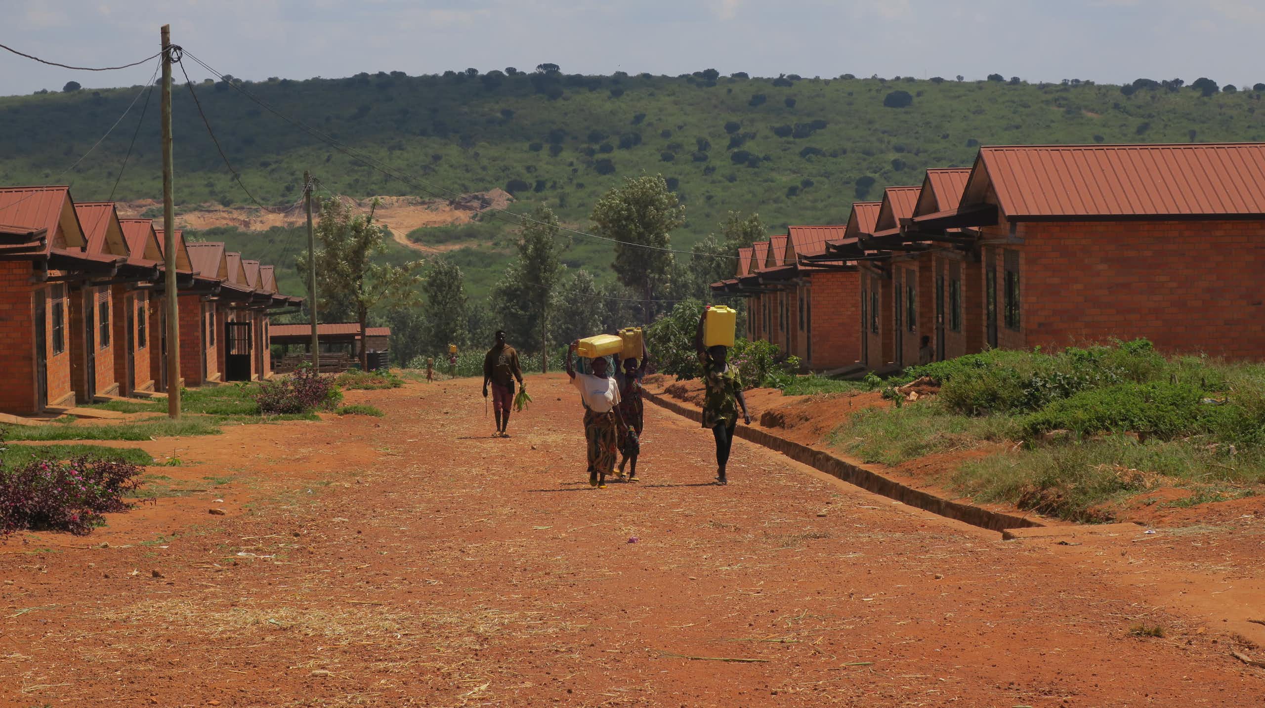 A dusty red road with two rows of houses on either side and people walking with 20 litre water buckets on their heads