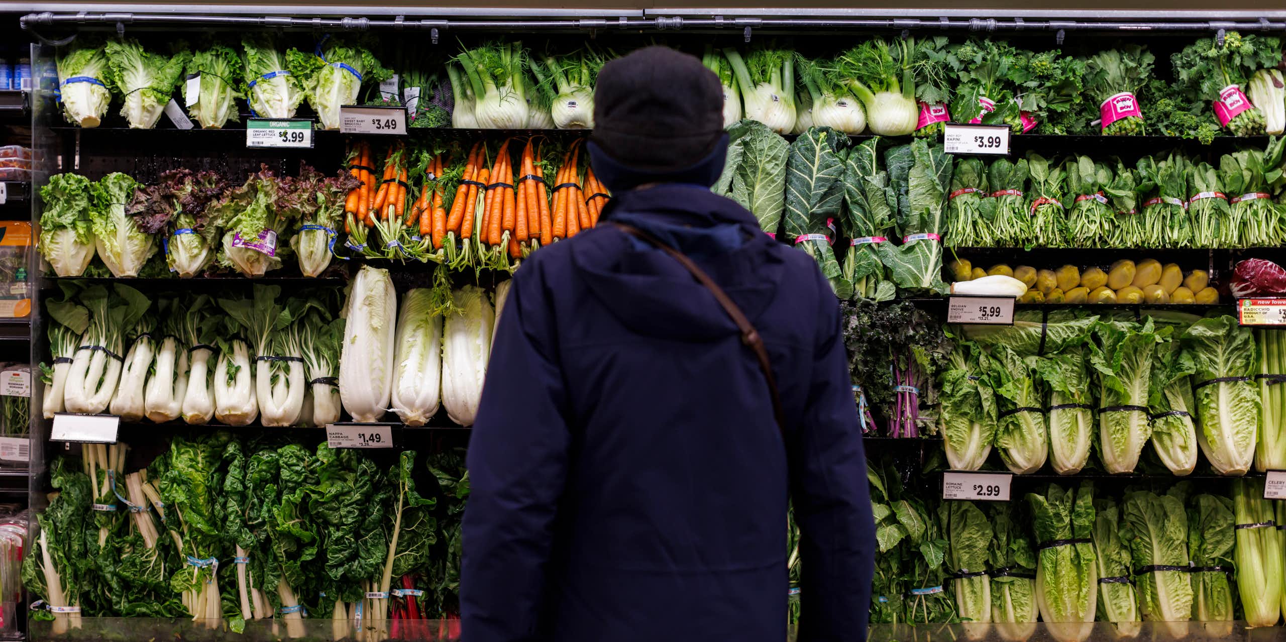 A person, seen from behind, stands in front of shelves of fresh vegetable produce in a grocery store