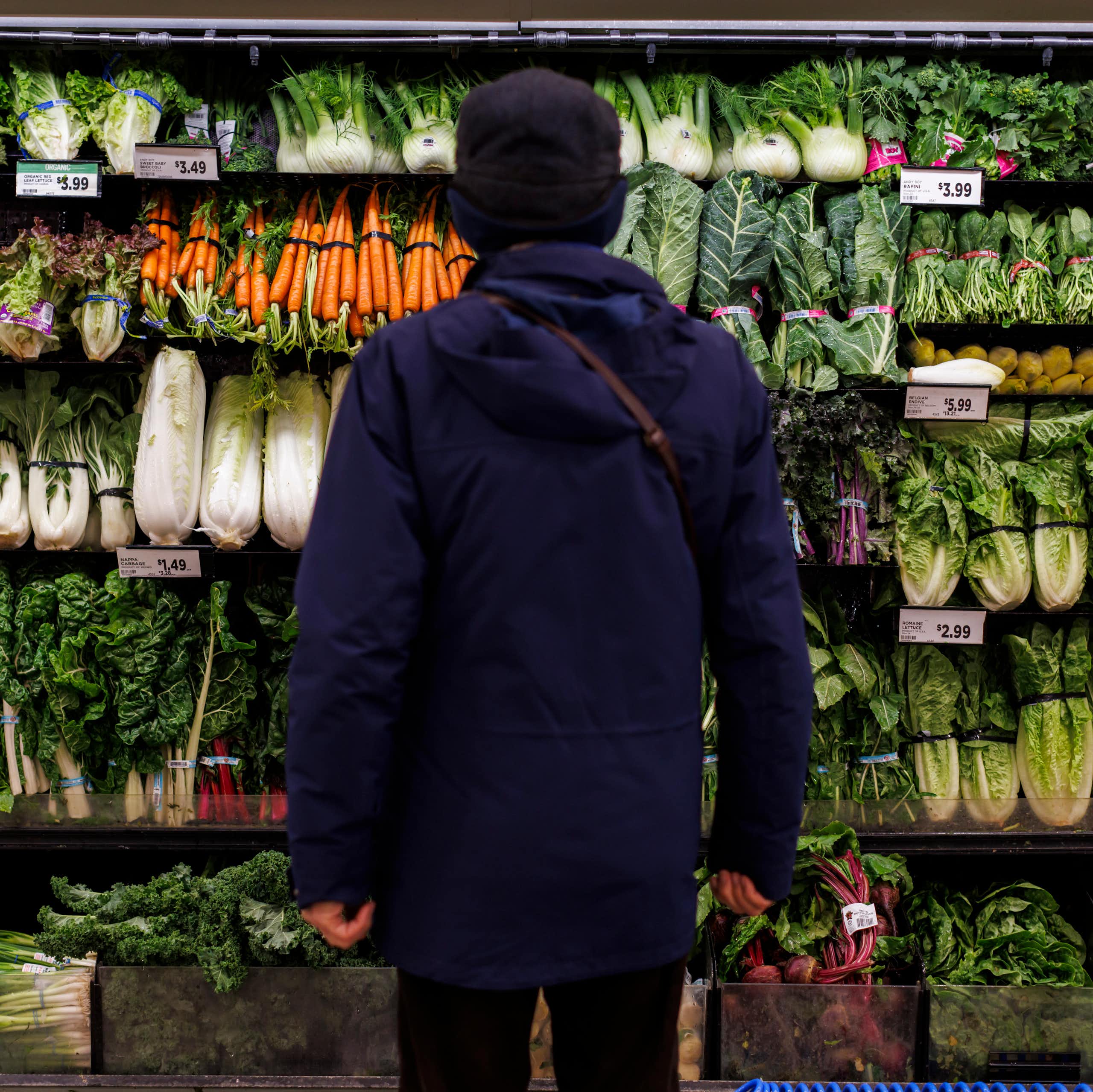 A person, seen from behind, stands in front of shelves of fresh vegetable produce in a grocery store