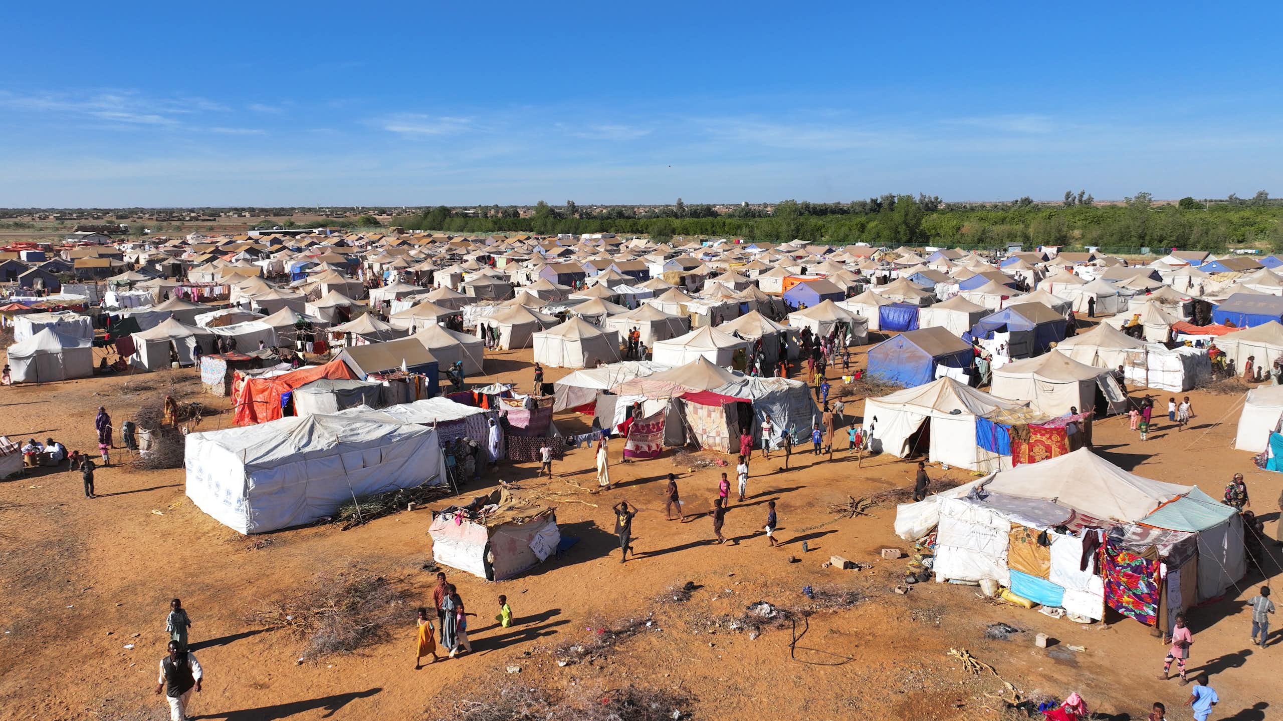 An aerial shot of a dusty field dotted with several white tents with several people walking around
