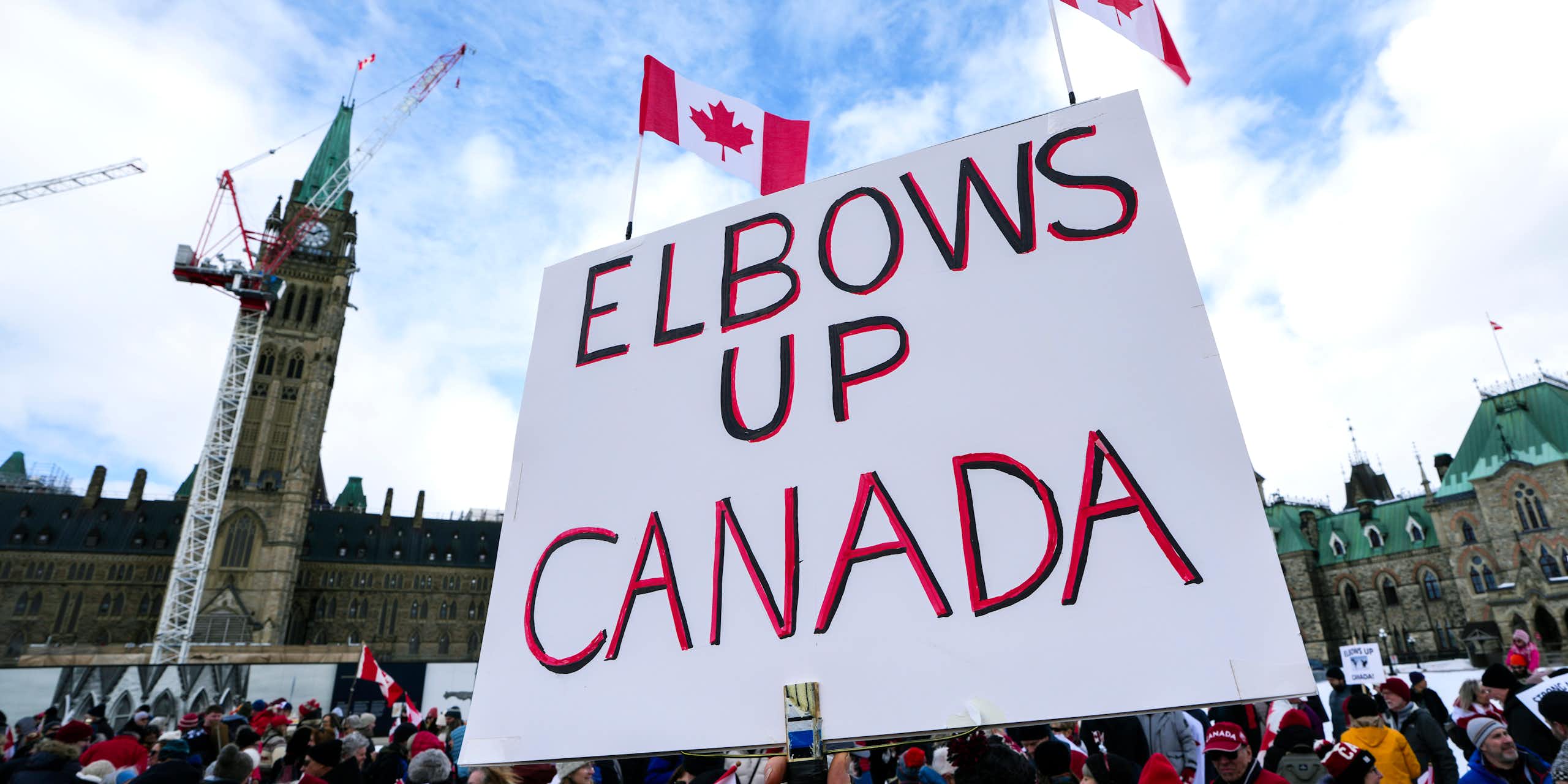 A person holding a white placard reading elbows up canada with two small canadian flags attached 