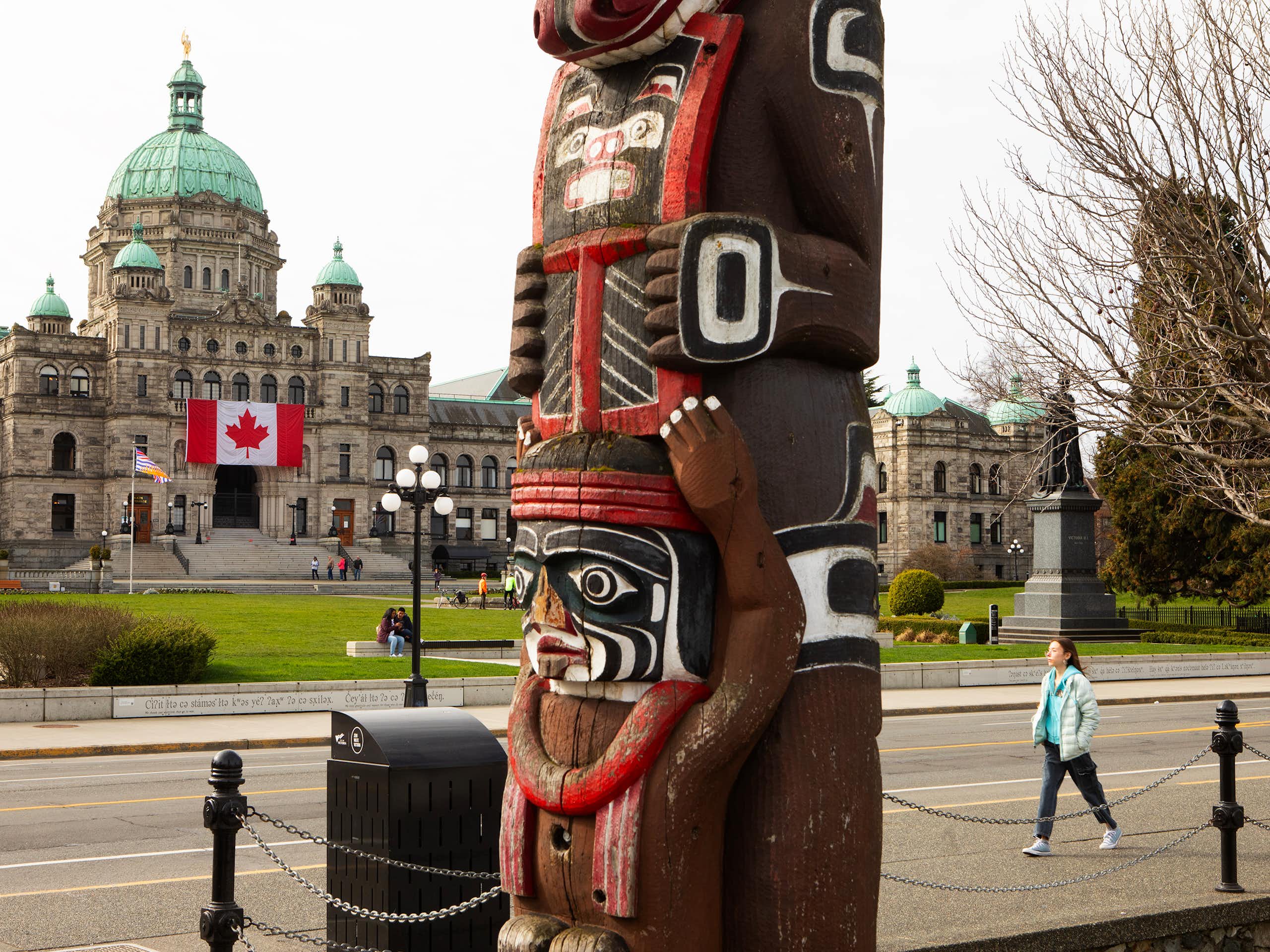 A totem pole is seen in the foreground, while a domed building with a Canadian flag over it is seen in the background