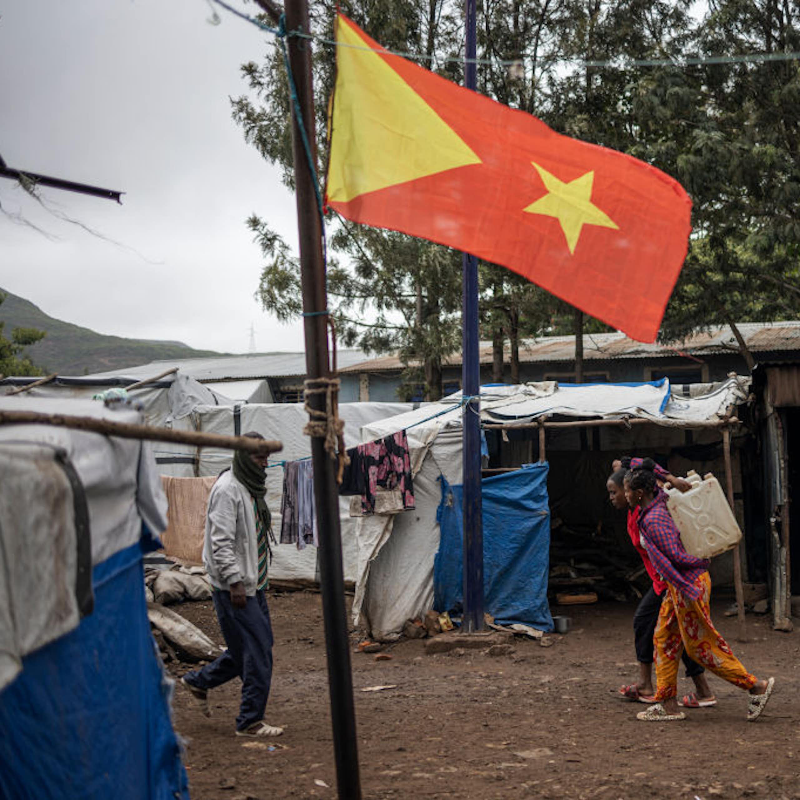 Three children and a man walking past makeshift shelters made with poles and white and blue canvas near a pole with a yellow and red flag