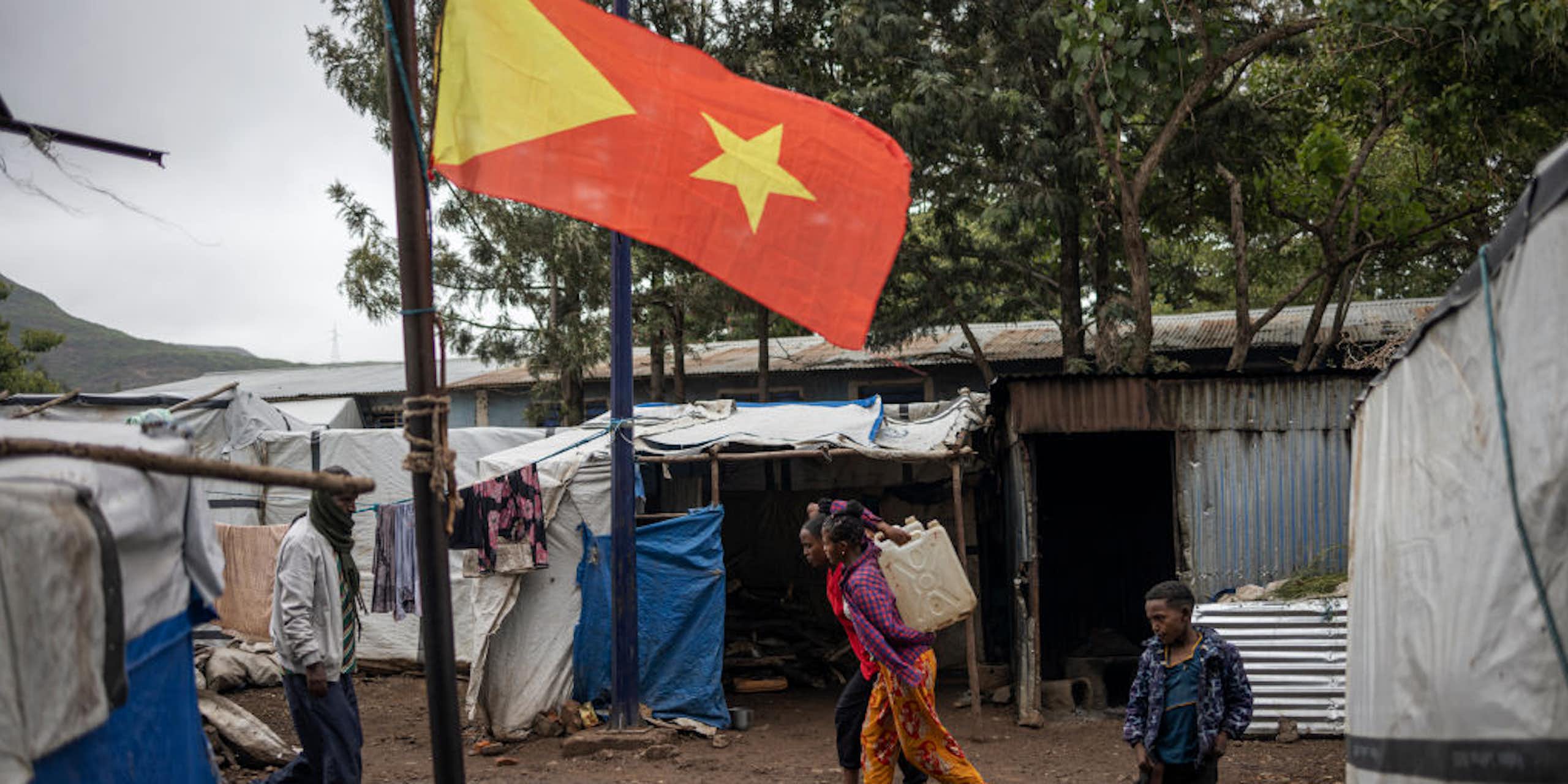 Three children and a man walking past makeshift shelters made with poles and white and blue canvas near a pole with a yellow and red flag