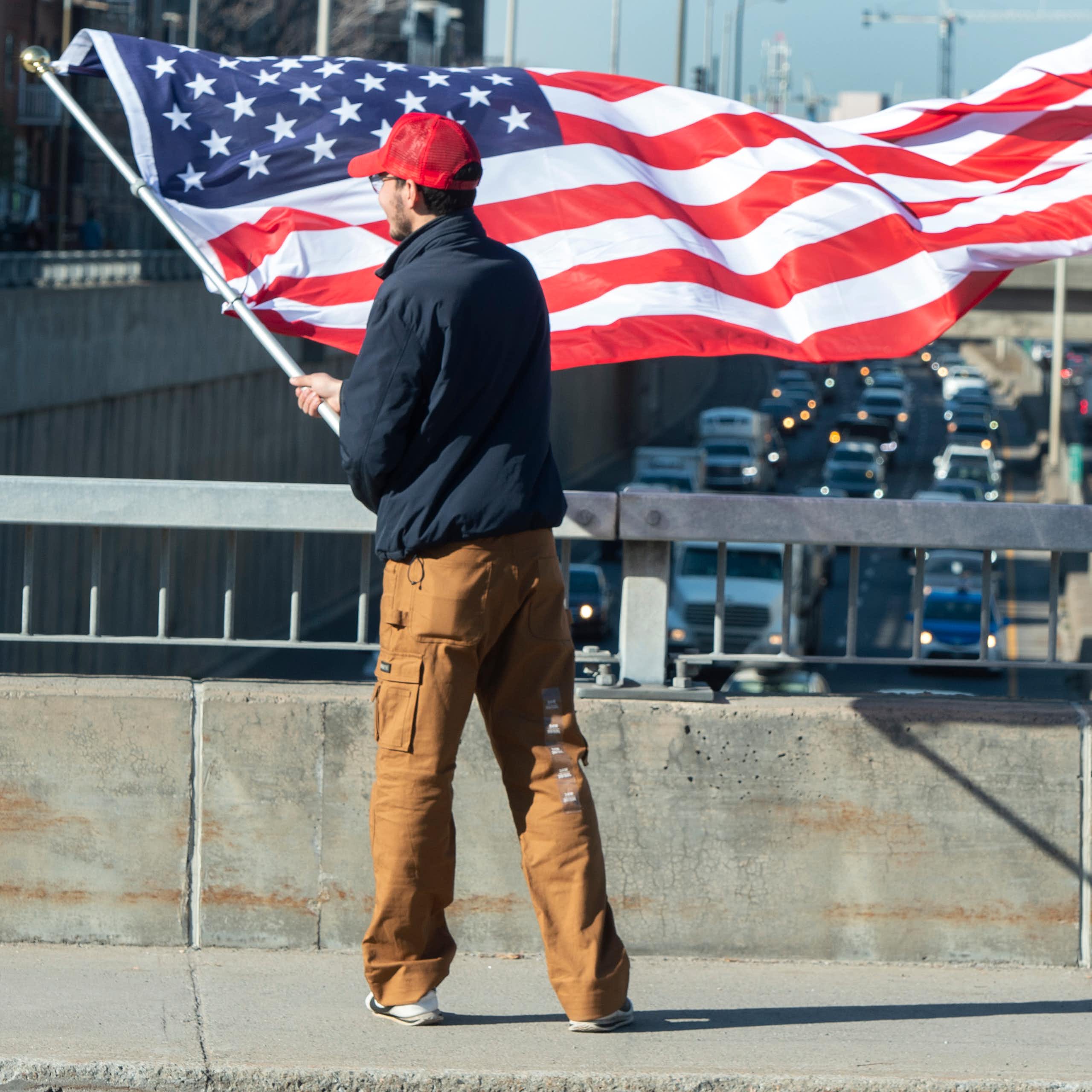 A man in a red ball cap is photographed from behind waving an American flag from an overpass.