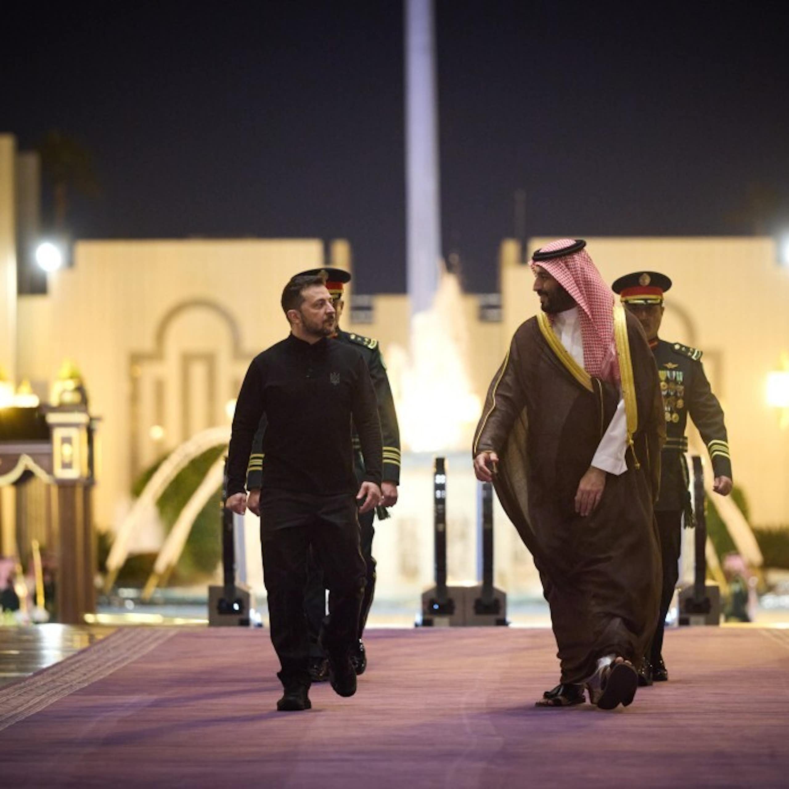 Two men walk on a carpet in front of an illuminated building..