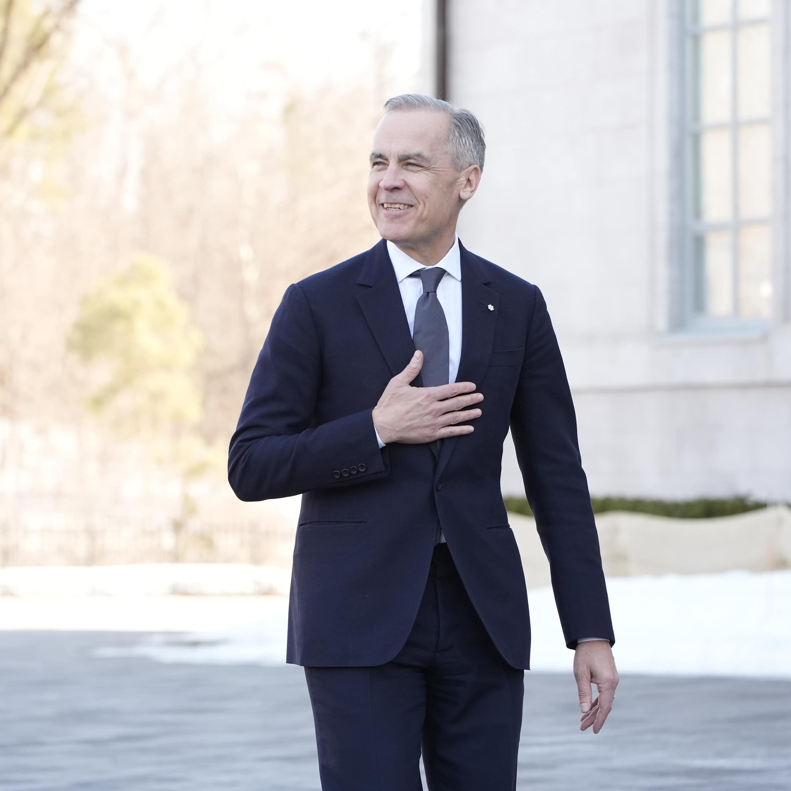 A thin man with grey hair in a dark suit smiles while walking outdoors, with his right hand on his chest.