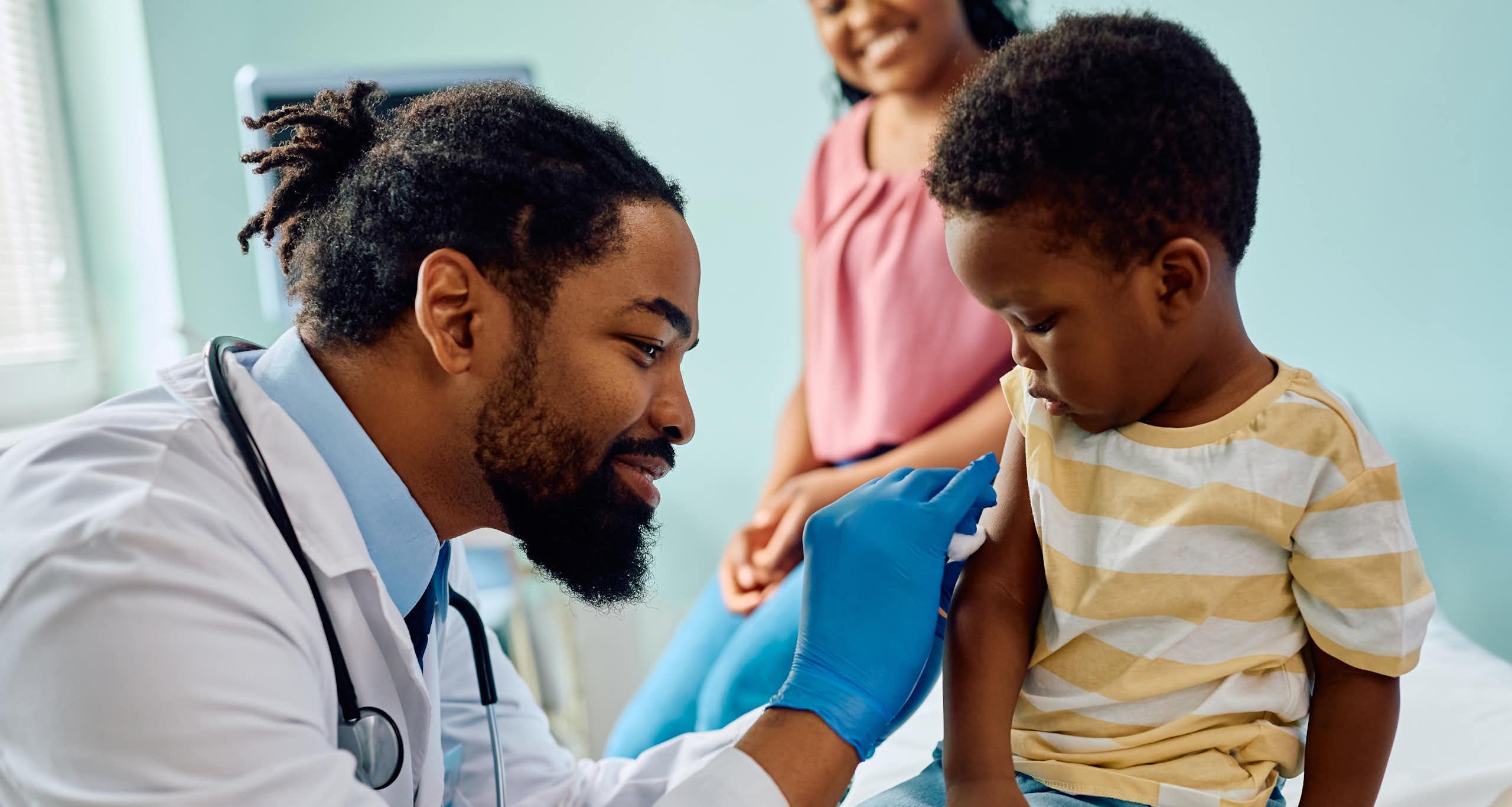 A doctor preparing a child's arm for an injection, with a parent in the background