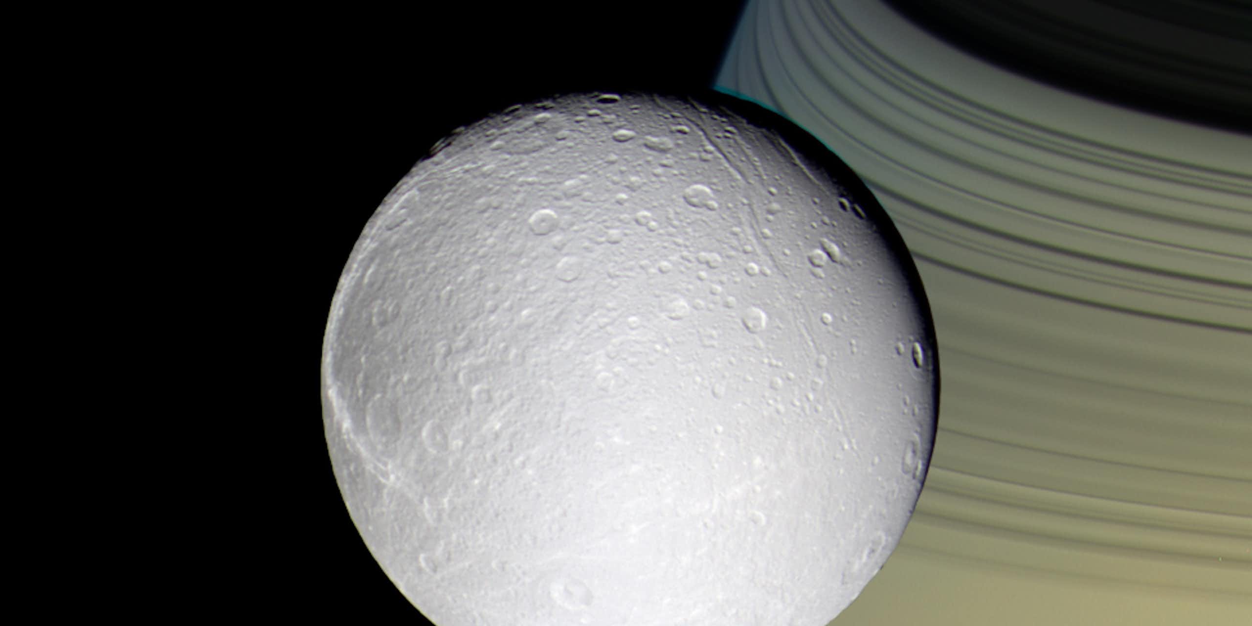 Foto de uma lua gelada com um planeta amarelado e anéis atrás,