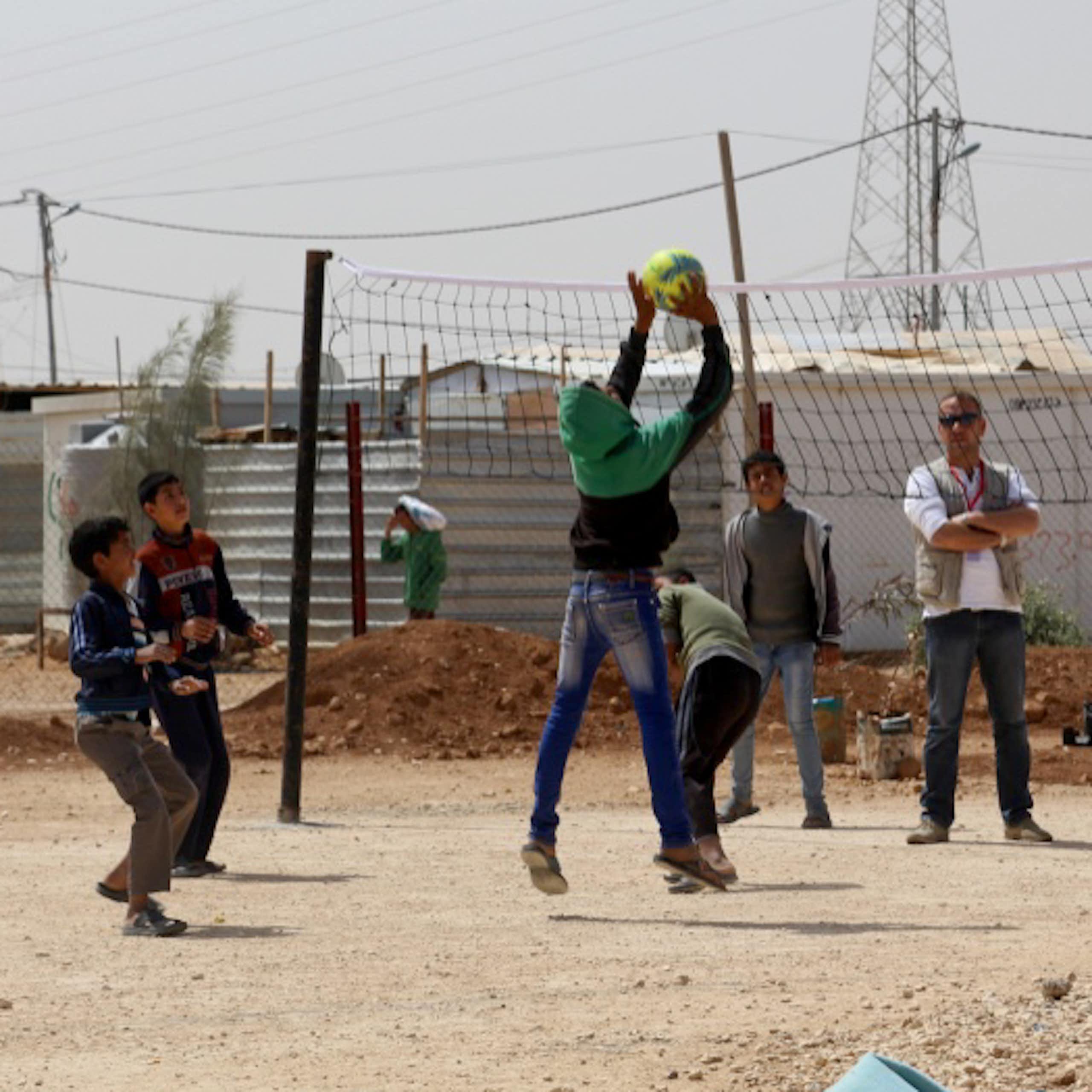 Group of young boys play basketball with refugee camp in background.