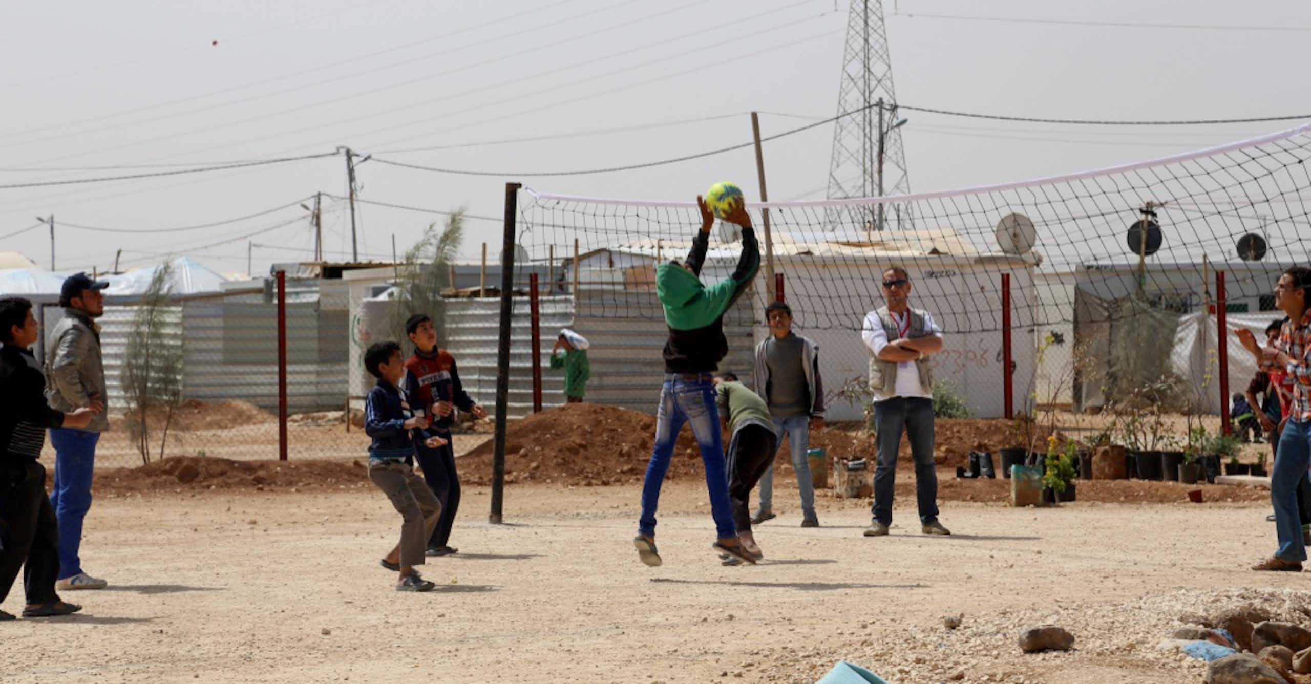 Group of young boys play basketball with refugee camp in background.
