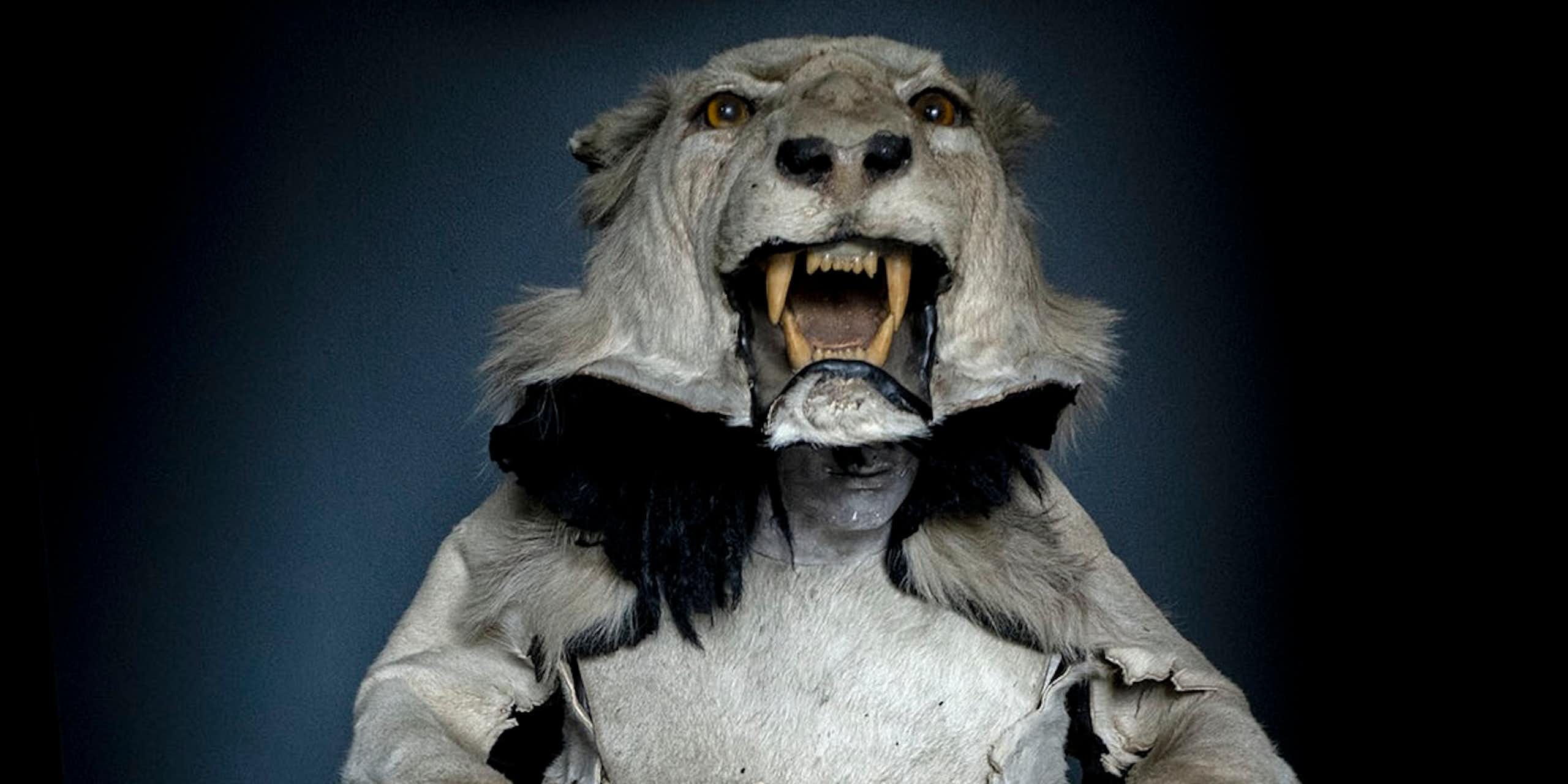 A mannequin wearing a stuffed lion's head as a hat. The lion holds two bags, each with a humanlike head in them.