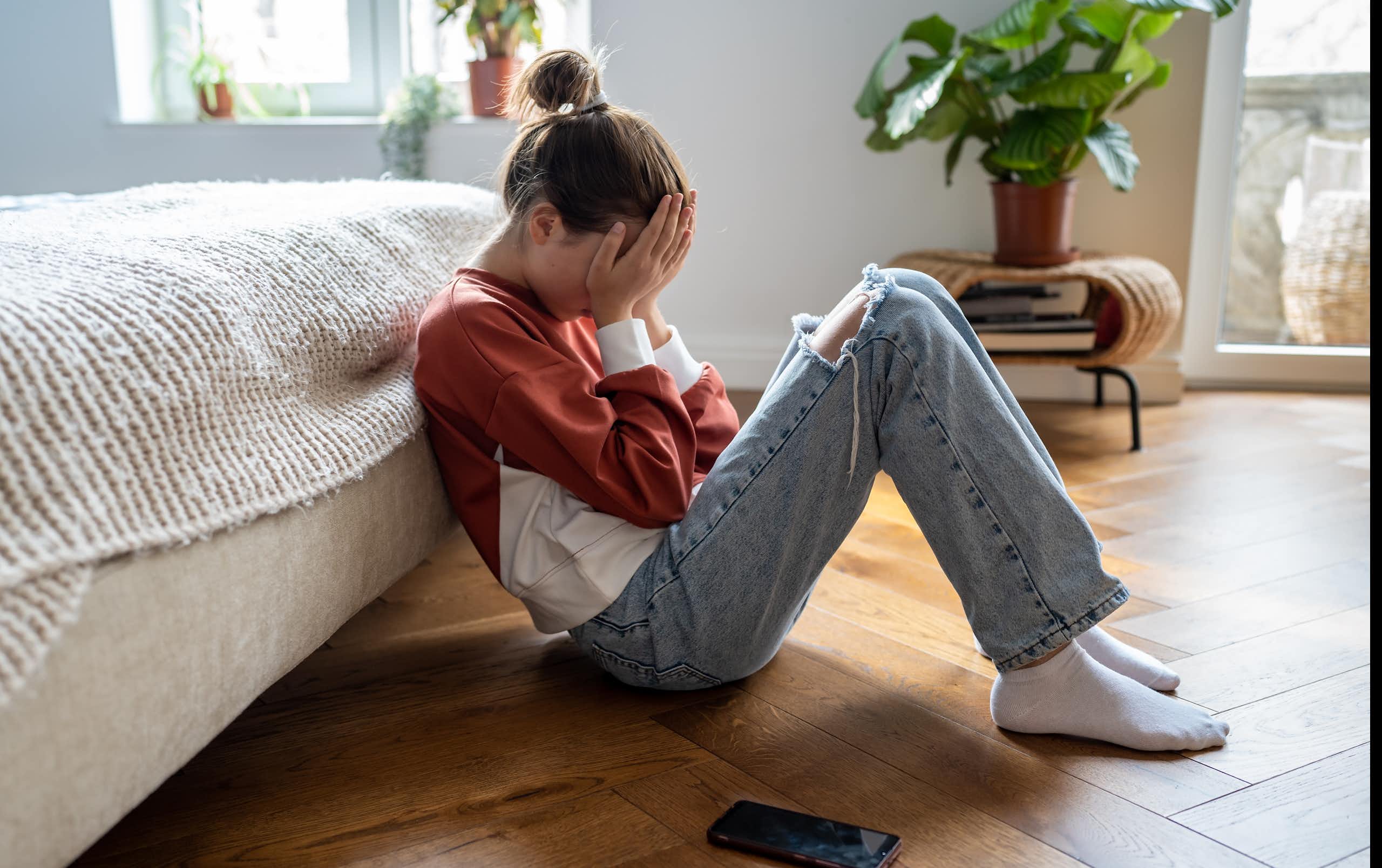 A girl sits on the floor leaning against a bed with her face in her palms