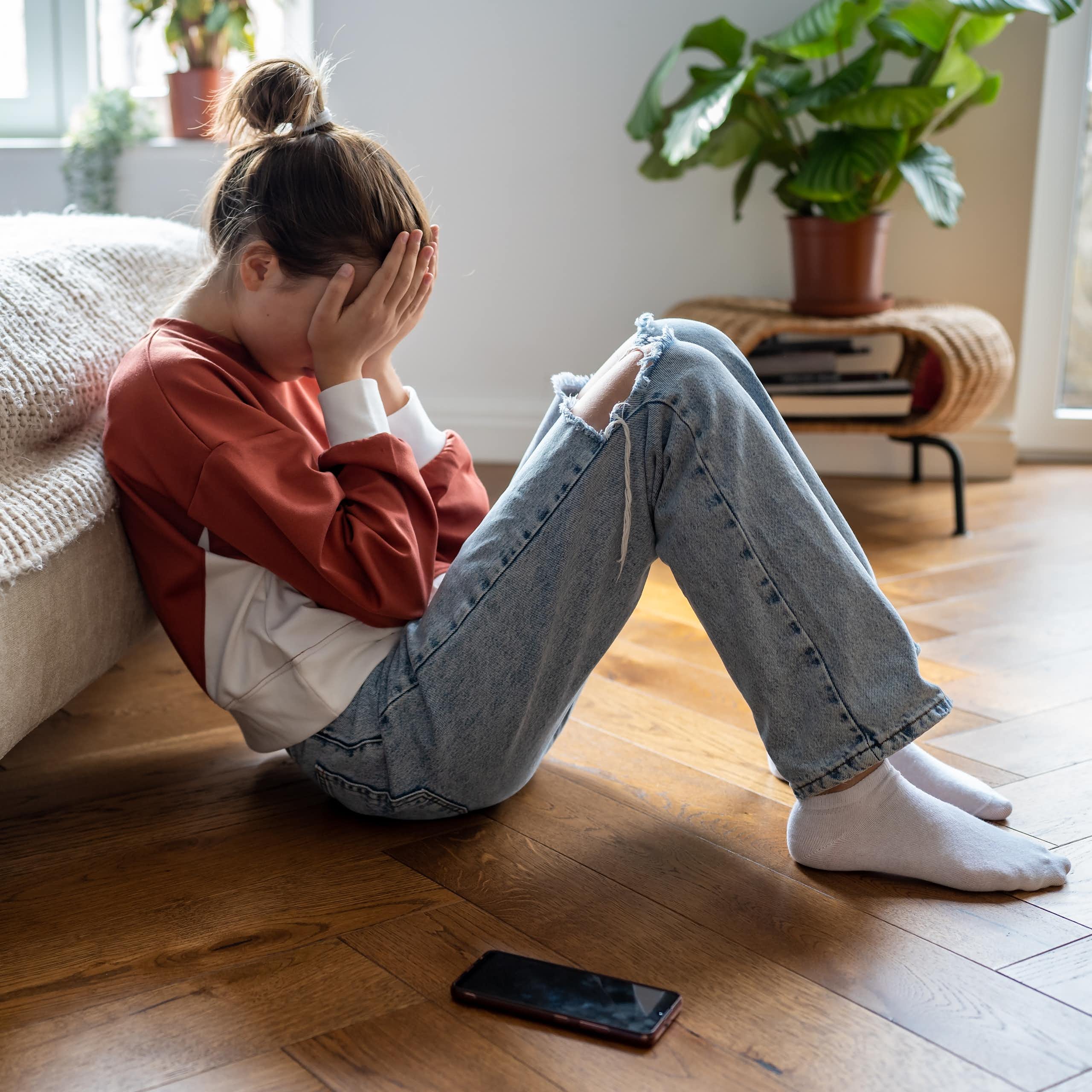 A girl sits on the floor leaning against a bed with her face in her palms